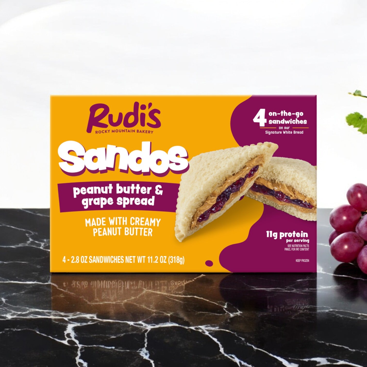 A box of Rudi's Rocky Mountain Bakery's Peanut Butter and Grape Spread Sandos, 11.2 oz - 1 Pack, sits on the table, presenting an ideal vegan choice. Fresh grapes are artfully arranged next to the box, enhancing its allure.