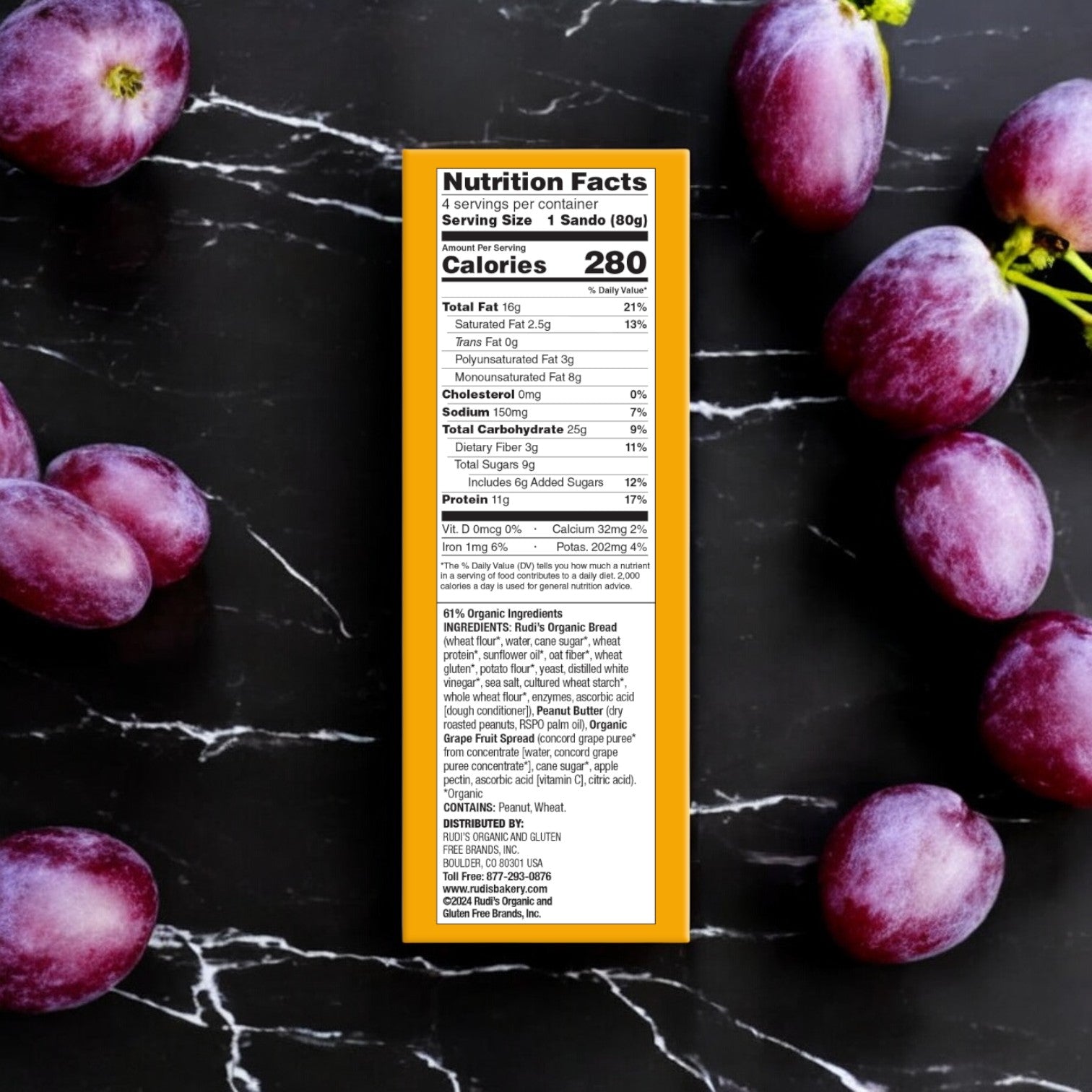 A nutrition facts panel of Rudi's Peanut Butter and Grape Spread Sandos, a vegan option from Rudi's Rocky Mountain Bakery, surrounded by red grapes on a sleek black marble surface.