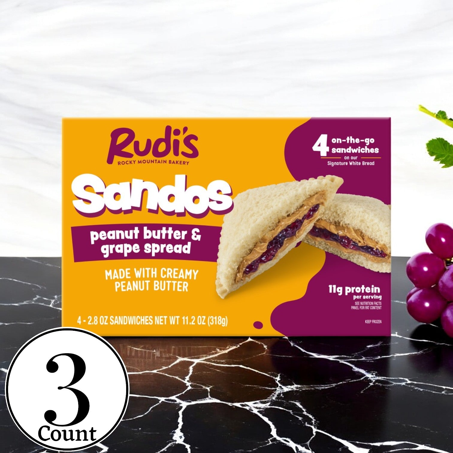 A box of Rudi's Peanut Butter and Grape Spread Sandos from Rudi's Rocky Mountain Bakery is placed on a wooden table, showcasing the vegan option. The packaging indicates it contains three packs and provides 11g of protein per serving. Grapes are visible beside the box.