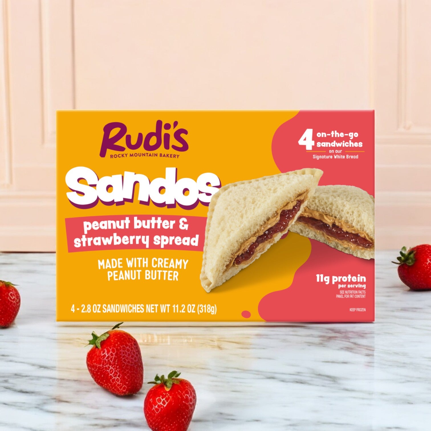 A pack of Rudi's Rocky Mountain Bakery Peanut Butter and Strawberry Spread Sandos, 11.2 oz, rests on a marble surface surrounded by fresh strawberries, providing a delightful vegan snack experience.