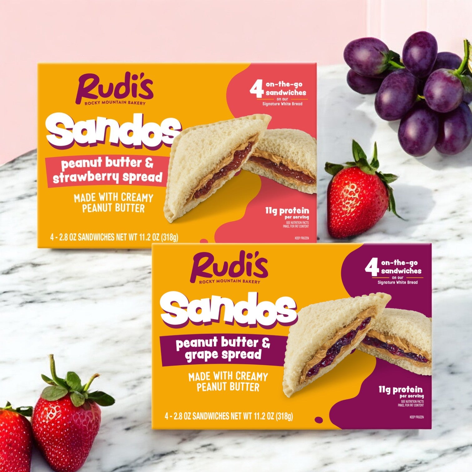 Two packs of Rudi's Variety Pack by Rudi's Rocky Mountain Bakery, featuring one Peanut Butter and Strawberry Spread Sandos and one Peanut Butter and Grape Spread Sandos. One pack is open, showcasing a delicious sandwich with fresh grapes and strawberries in the background, emphasizing these tasty vegan snacks.