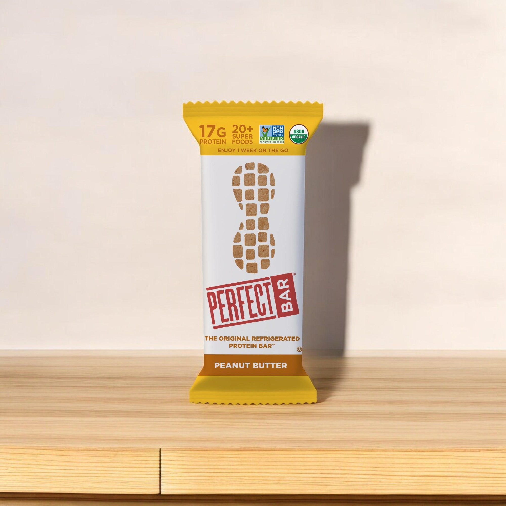 A Perfect Bar Gluten-Free Peanut Butter Refrigerated Protein Bar, 2.3 oz - 1 Count, is positioned on a wooden surface against a plain background. The wrapper emphasizes its 17g protein content and inclusion of various organic superfoods.