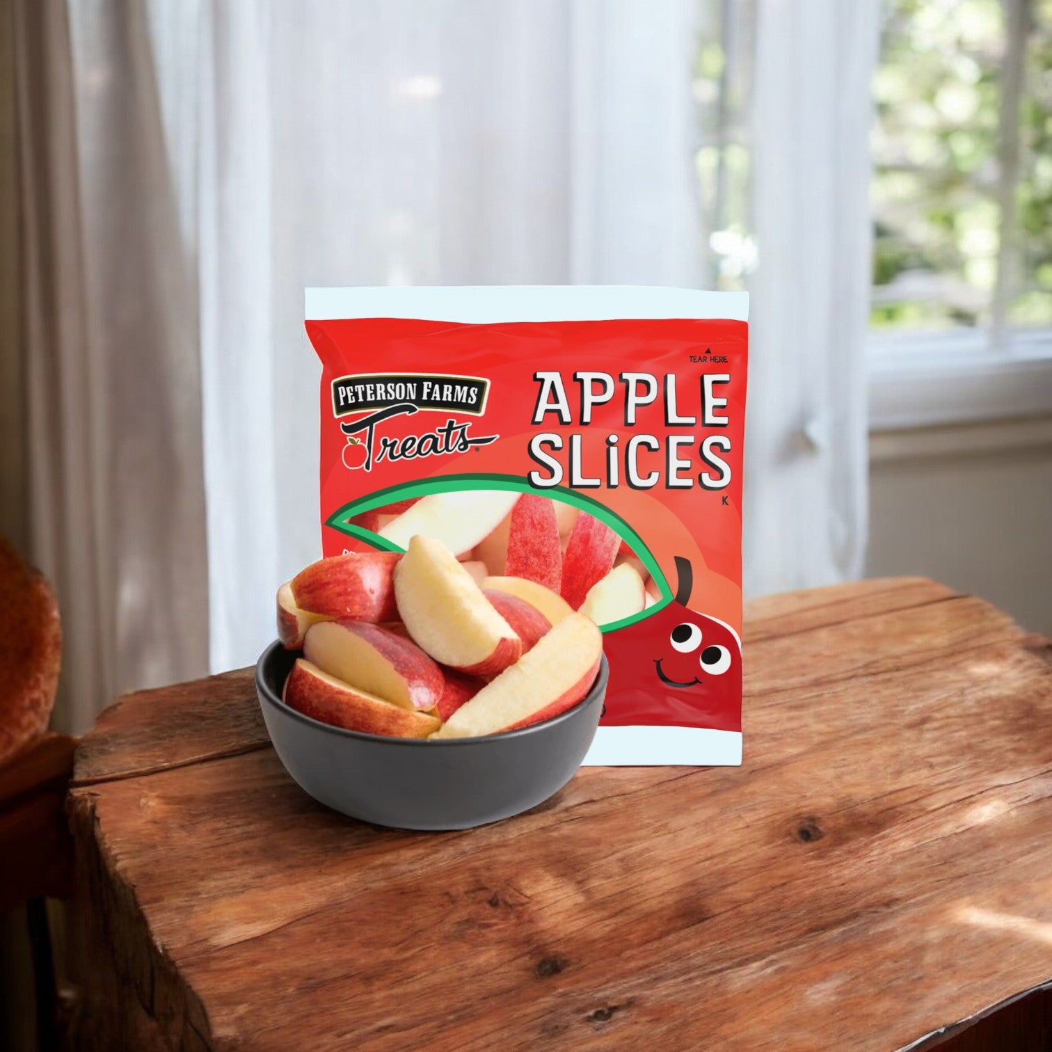 A bowl of fresh apple slices sits on a wooden table with a bag labeled "Peterson Farms Fresh Sliced Apple Packs for Convenient Snacking" behind it, highlighting the ideal healthy snack. White-curtained windows are in the background.