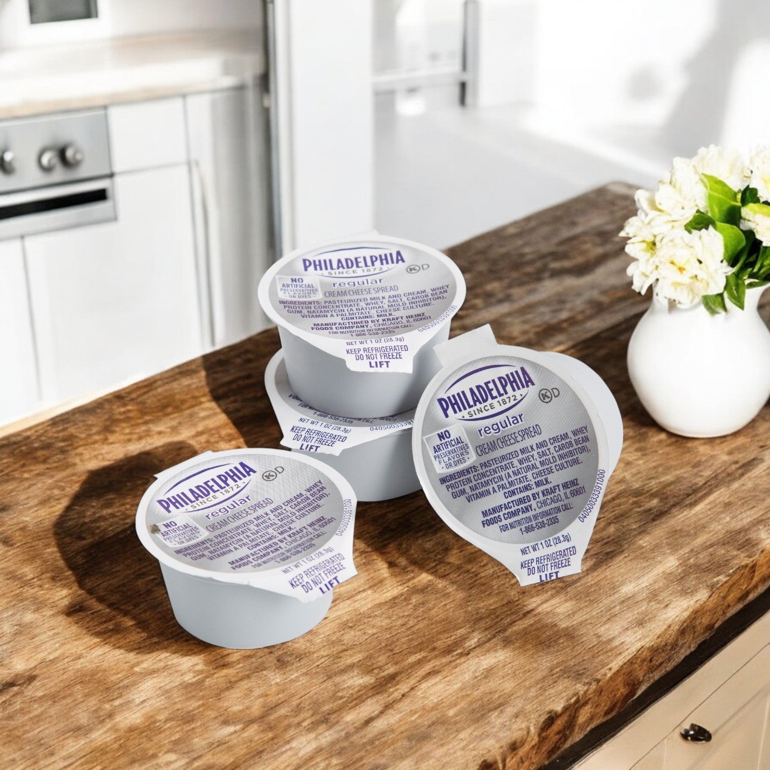 Four small cups of Kraft Philadelphia Cream Cheese Original Spread sit on a wooden kitchen countertop near a white vase of flowers.