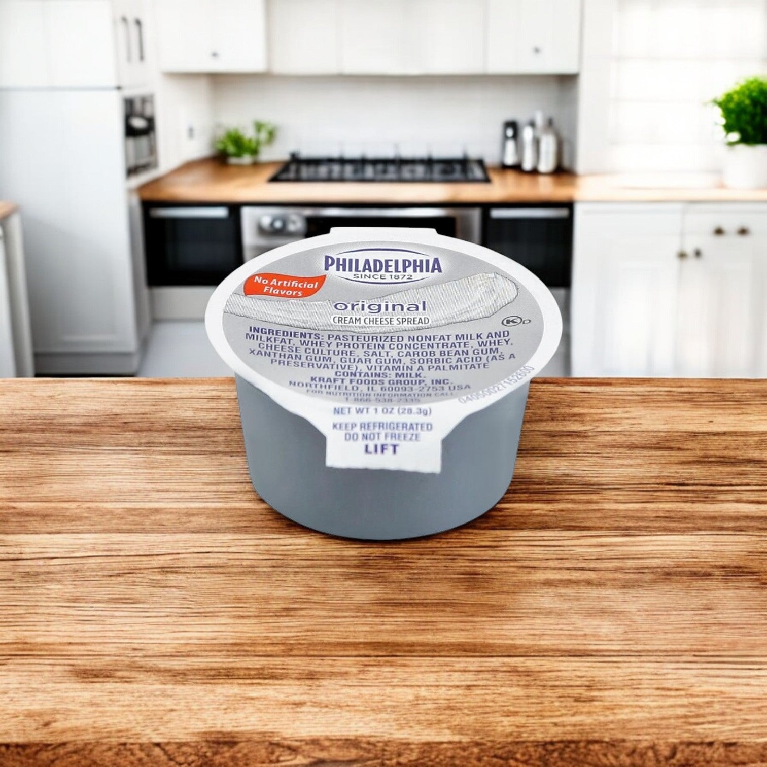 A 1 oz. cup of Kraft's Philadelphia Cream Cheese Original Spread rests on a wooden kitchen counter, with a blurred white kitchen in the background enhancing this creamy delight's appeal.
