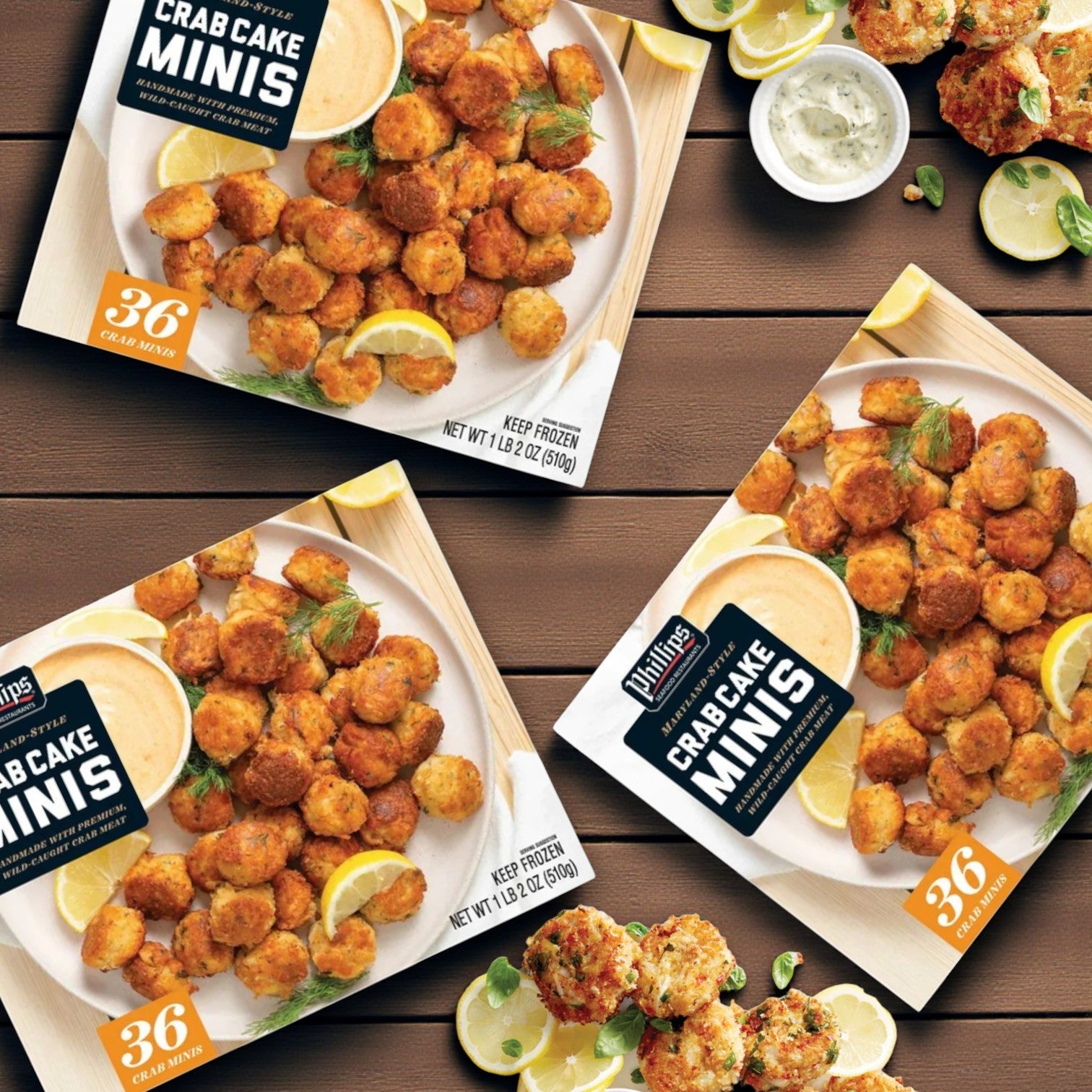 Three boxes of Phillips Crab Cake Minis – 1 lb 2 oz, 3 Pack (108 count) by Phillips Foods are displayed on a wooden surface with sauce, lemon wedges, and herbs—an enticing seafood appetizer spread.