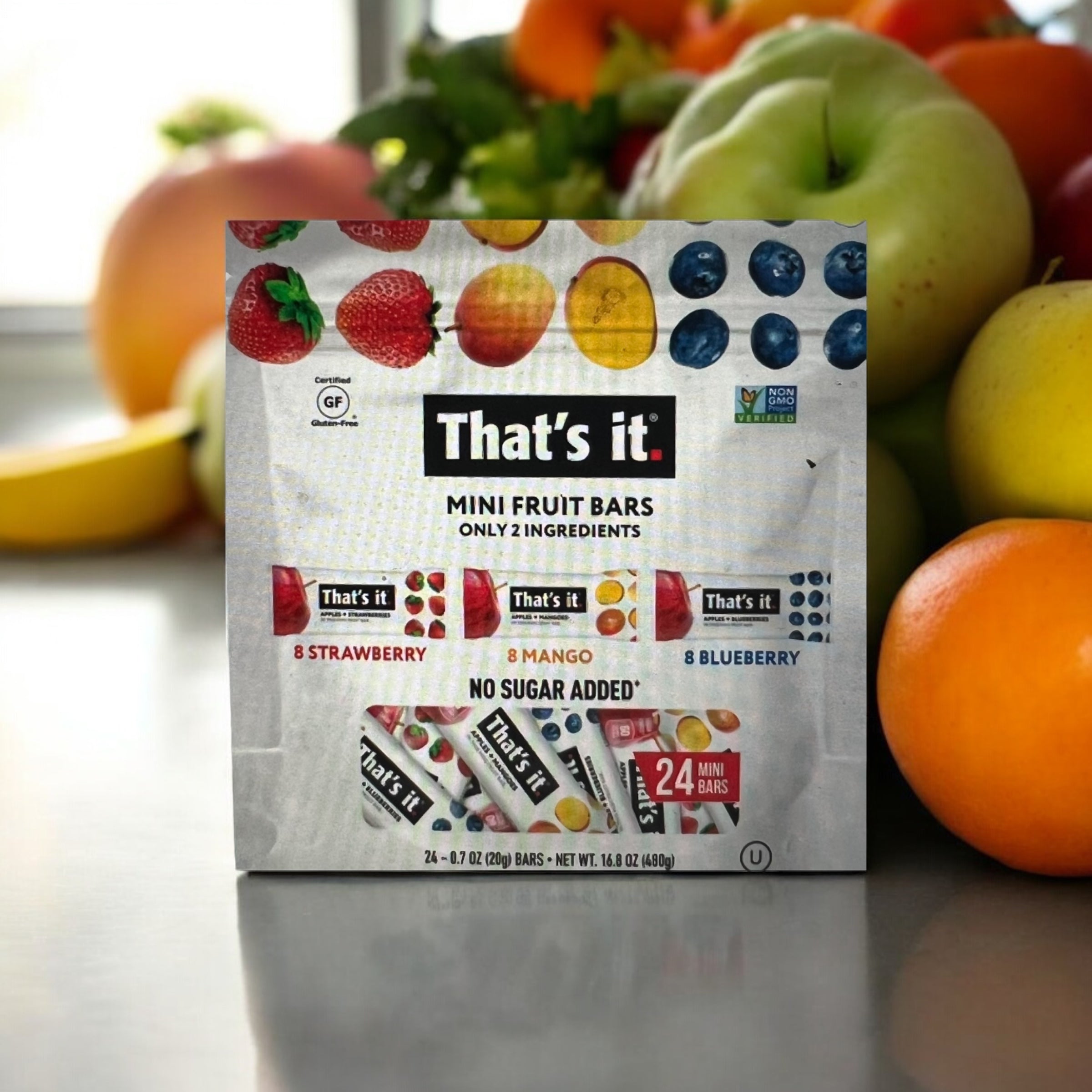 A package of "That’s it Mini Fruit Bars" from Easy Lunches is displayed, featuring 24 bars with no sugar added. The flavors included are 8 strawberry, 8 mango, and 8 blueberry. Fresh fruits are softly blurred in the background.