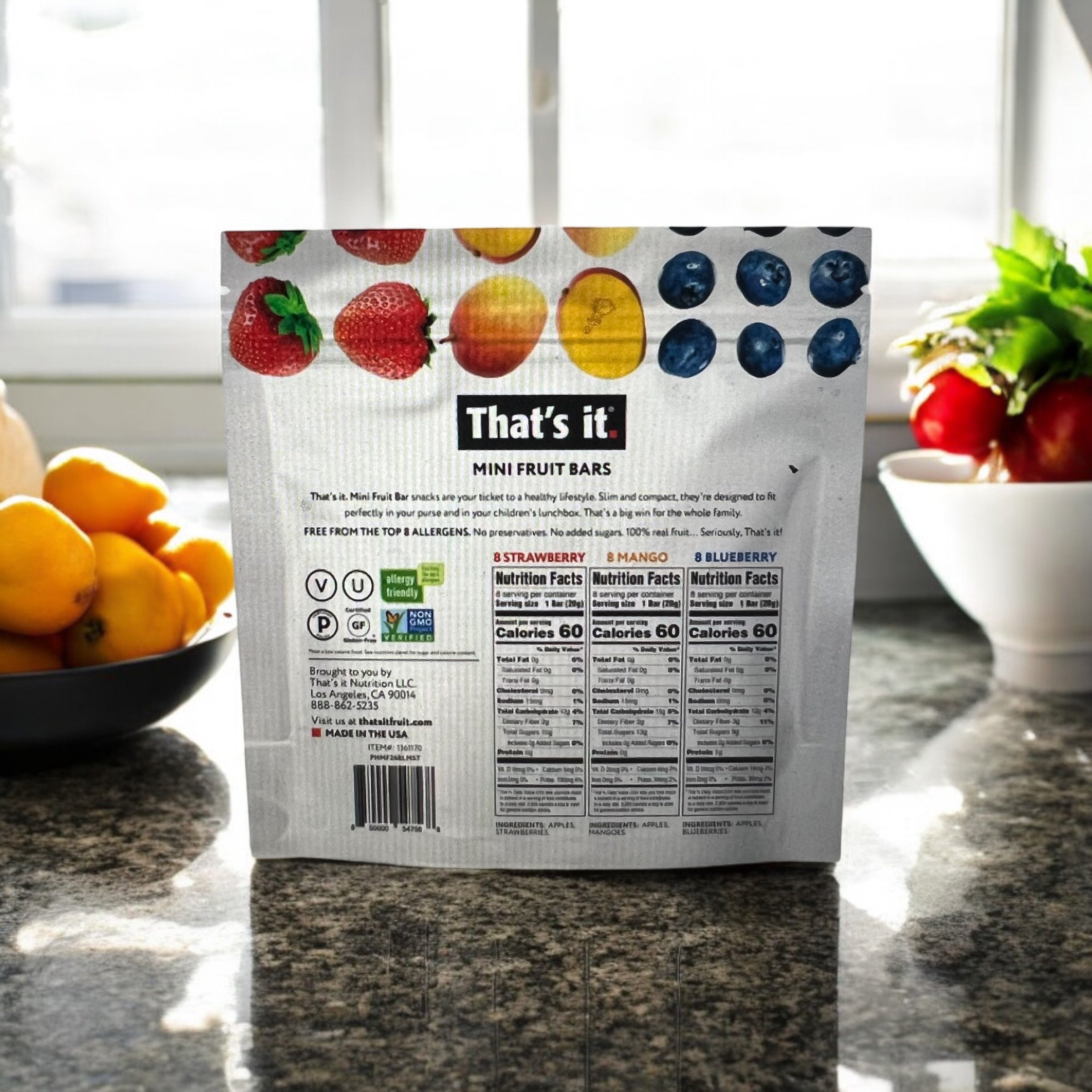 A package of Easy Lunches That’s it Mini Fruit Bars, Strawberry, Mango, Blueberry Flavor (24 count), displaying the nutritional information and images of strawberries, mangoes, and blueberries. The package is placed on a kitchen counter with a bowl of oranges nearby.