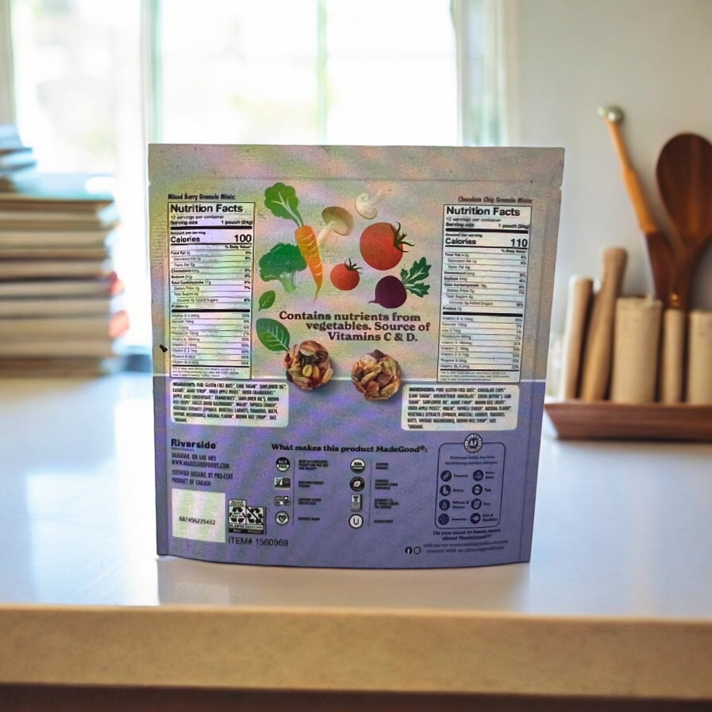 A back view of a packaged food product showcases the nutrition facts and ingredients through a window, situated near some books and kitchen utensils. This allergen-free, healthy snack from Easy Lunches – the MadeGood Granola Minis, Variety Pack (24 count) – is perfect for those mindful of their diet.