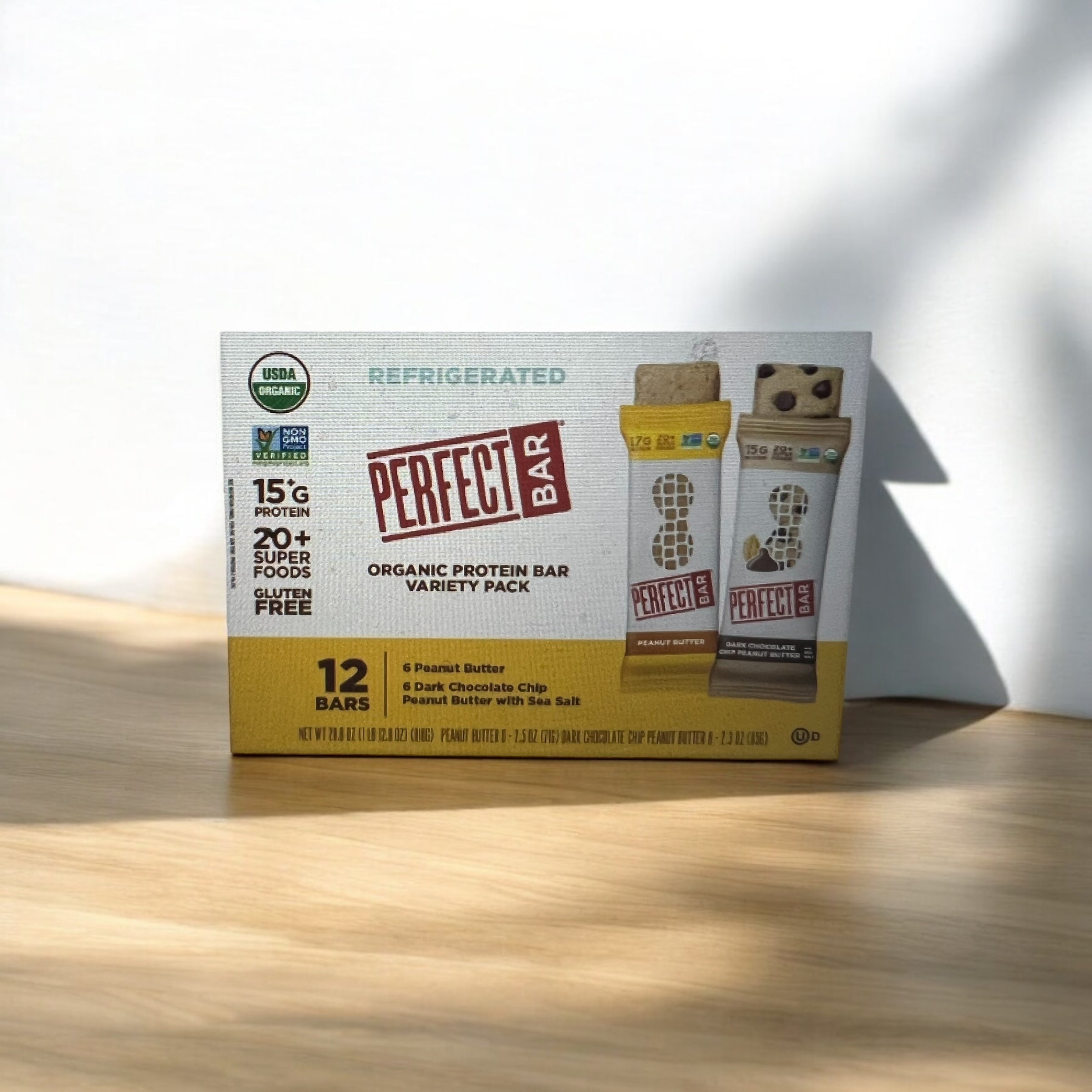 A box of Easy Lunches Perfect Bar Refrigerated Organic Protein Bars sits on a wooden surface in sunlight. The box mentions 12 bars, with flavors including Peanut Butter and Dark Chocolate Chip, each containing 15g of protein.