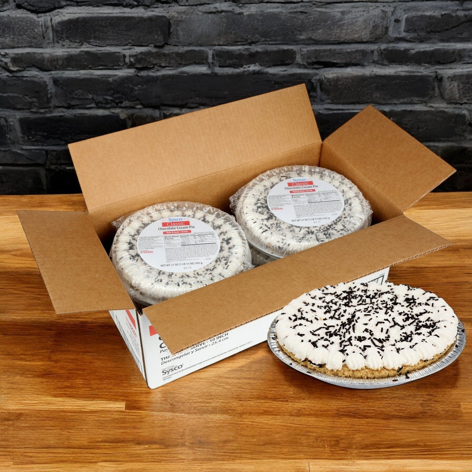 A Sysco Classic cardboard box with two sealed Chocolate Cream Pies sits beside an unwrapped pie on a wooden table. Each 10" pie features chocolate sprinkles and rich chocolate filling, ready to thaw and serve, all beautifully positioned in front of a brick wall.