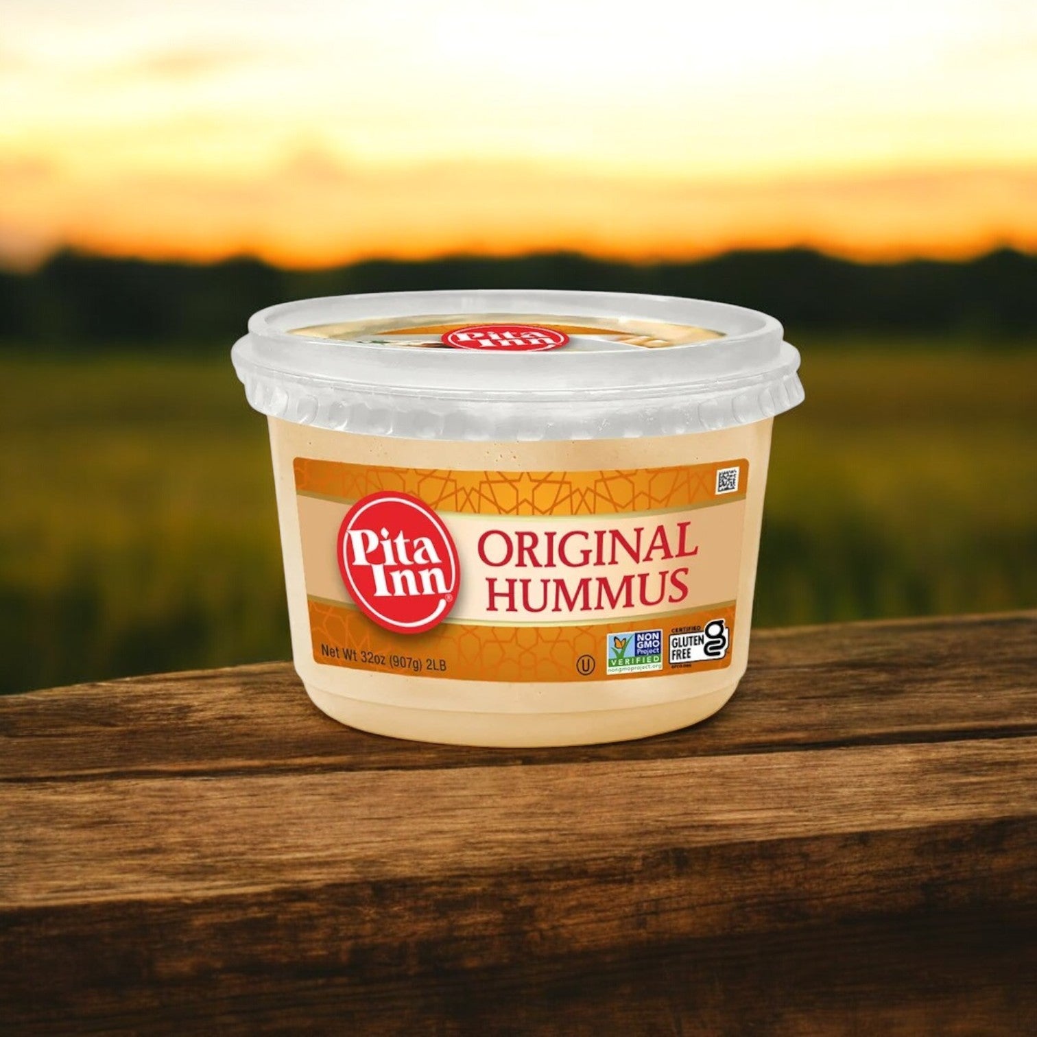 A 2 lbs. container of Pita Inn Original Hummus by Good Foods, known for its authentic Mediterranean flavor, rests on a wooden surface with a blurred outdoor background at sunset.