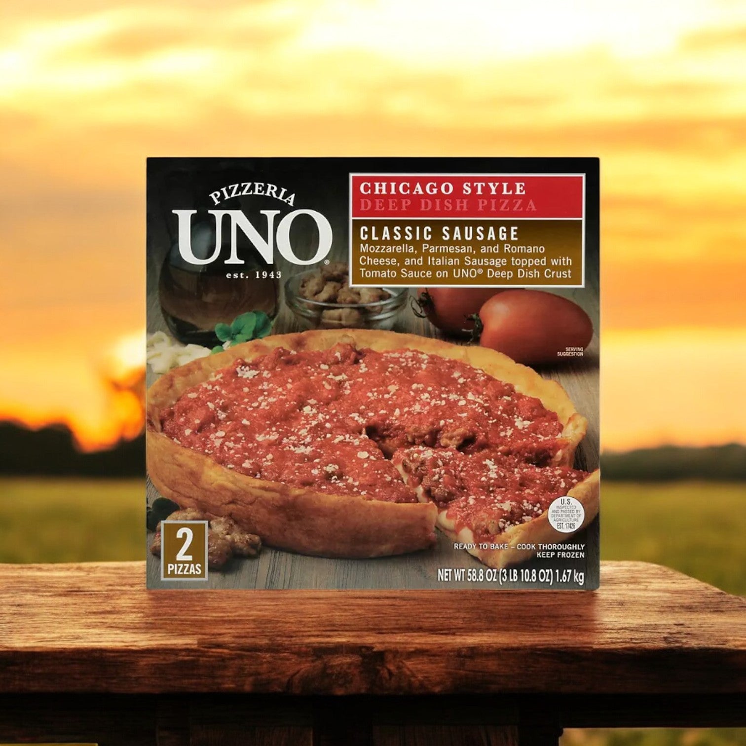 A case of Pizzeria Uno 9" Deep Dish Classic Sausage Pizza, featuring three packs with a total of six pizzas, resting on a wooden surface against a sunset backdrop.