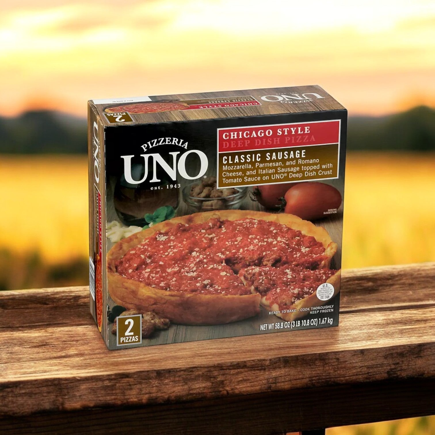 A case of Pizzeria Uno 9" Deep Dish Classic Sausage Pizza, consisting of 3 packs and a total of 6 pizzas, is placed on a wooden surface with a blurred outdoor background, embodying the essence of authentic Chicago-style pizza.