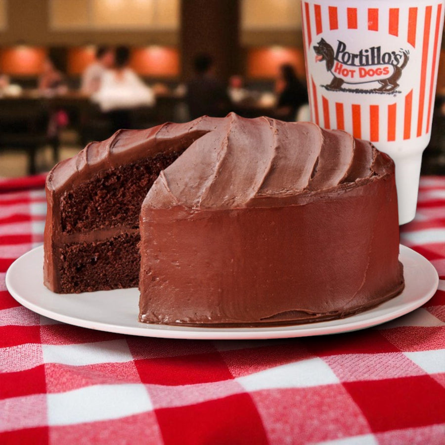 A slice of Portillo’s Famous Chocolate Cake! Two-Layer Delight by Portillo's rests on a white plate beside a Portillo's Hot Dogs cup atop a red and white checkered tablecloth. Ships frozen; serves 8-12 slices.