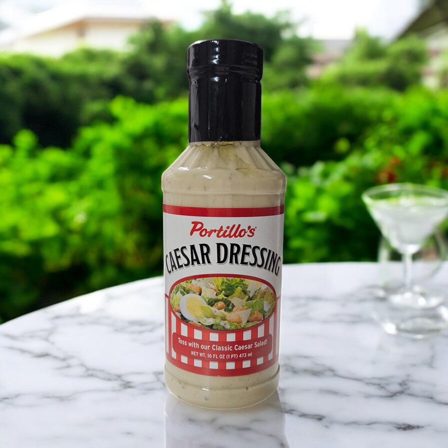 A bottle from the Portillo's 3-Pack Dressings set sits elegantly on a marble table with its label gleaming. In the blurred background, a lush garden and an inviting cocktail glass capture the essence of Portillo's.