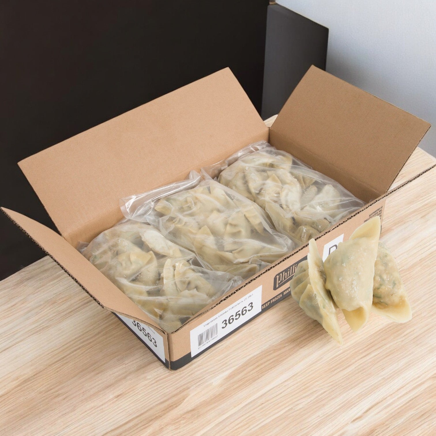 An open cardboard box from Phillips Foods rests on a wooden table, its contents revealing several sealed plastic bags of Potsticker Vegetable Shiitake Mushroom dumplings. A few dumplings are artfully arranged in front, suggesting an authentic flavor profile reminiscent of the delectable taste associated with Chesapeake cuisine.