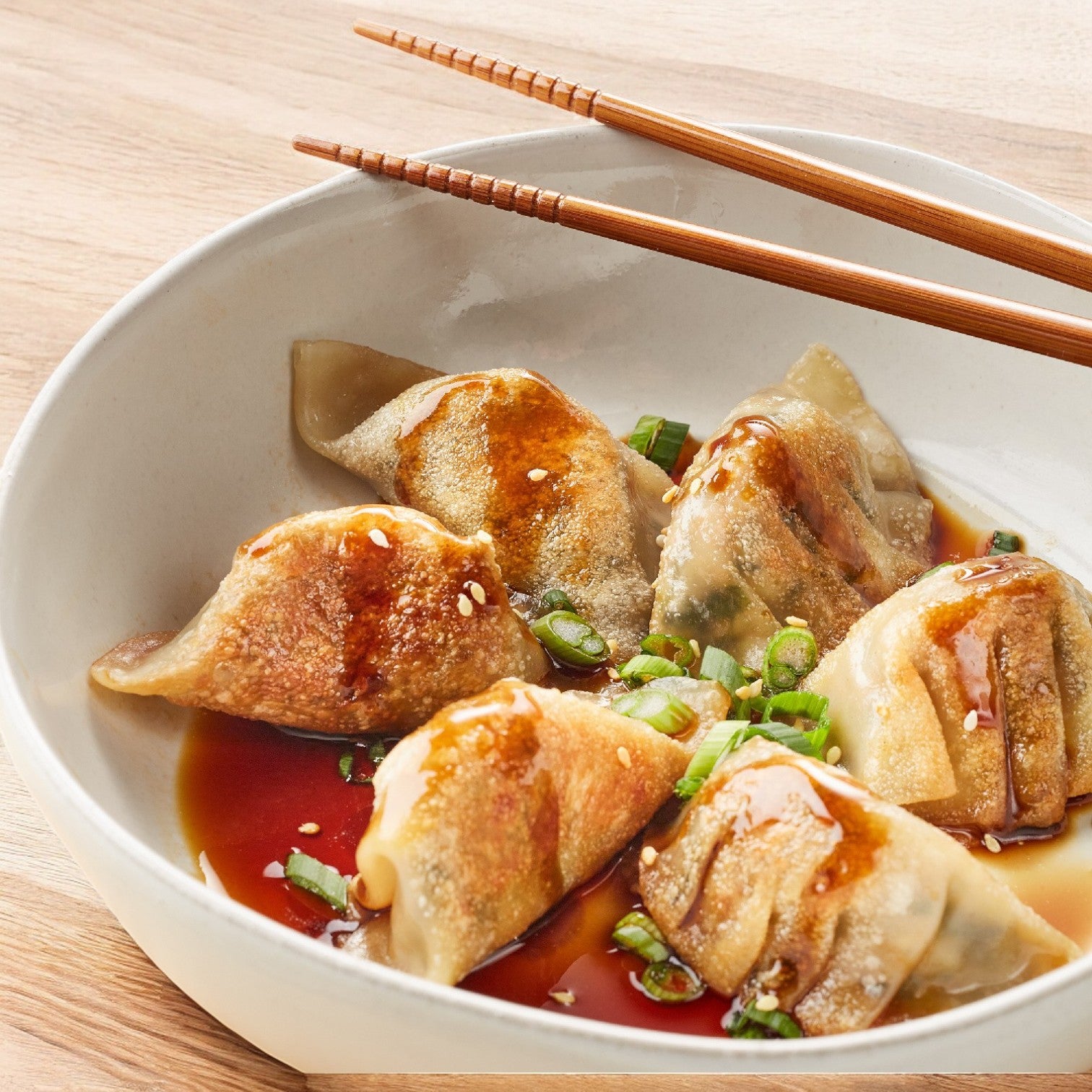 A bowl of Phillip Foods Potsticker Vegetable Shiitake Mushroom dumplings captures an authentic Chesapeake flavor, topped with soy sauce, sesame seeds, and chopped green onions. A pair of wooden chopsticks rests on the bowl's edge, ready to elevate this tasty fusion experience.