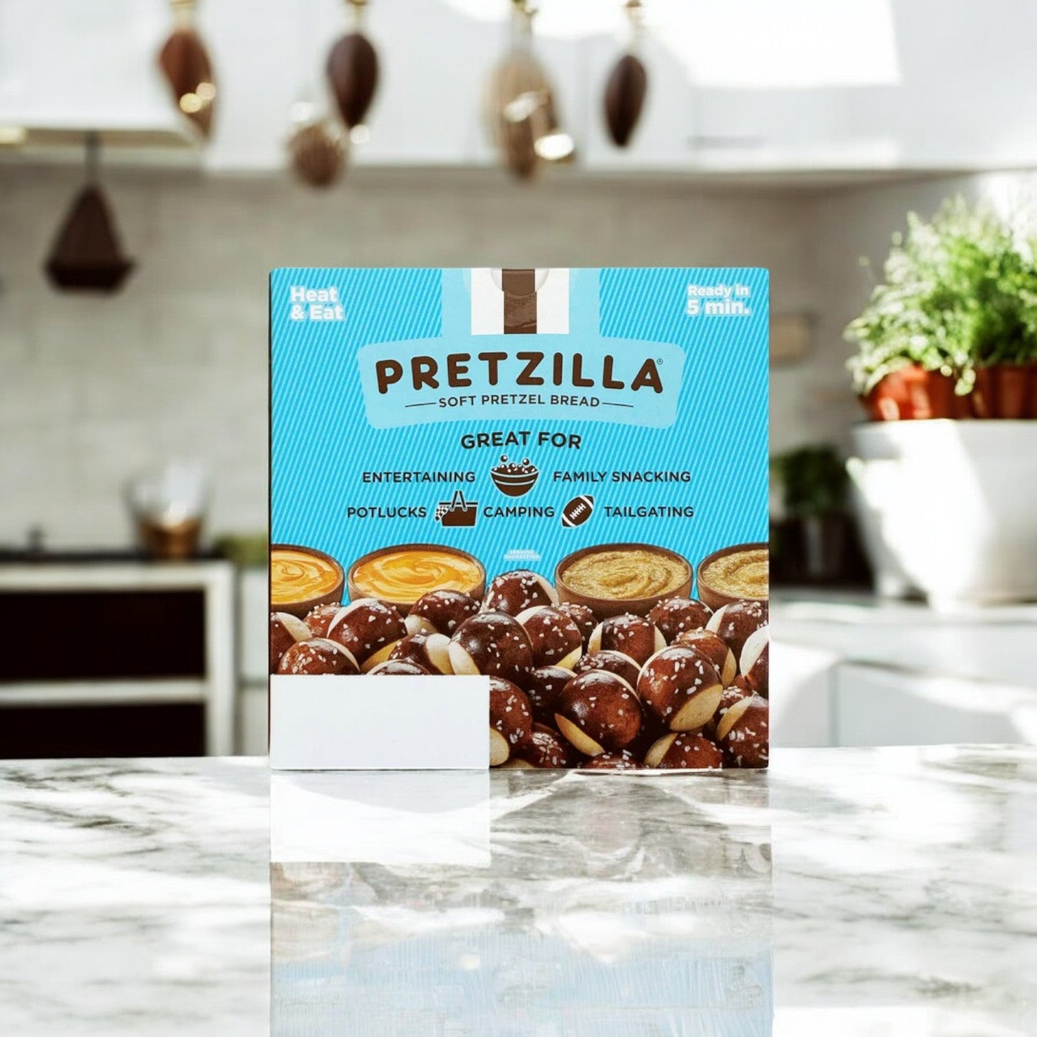 A 36.5 oz. box of Pretzilla Pretzel Bites sits invitingly on a marble kitchen counter, ready to satisfy snack cravings. The bright kitchen, adorned with hanging utensils and potted plants, emphasizes the convenience of its resealable bag for maintaining freshness.