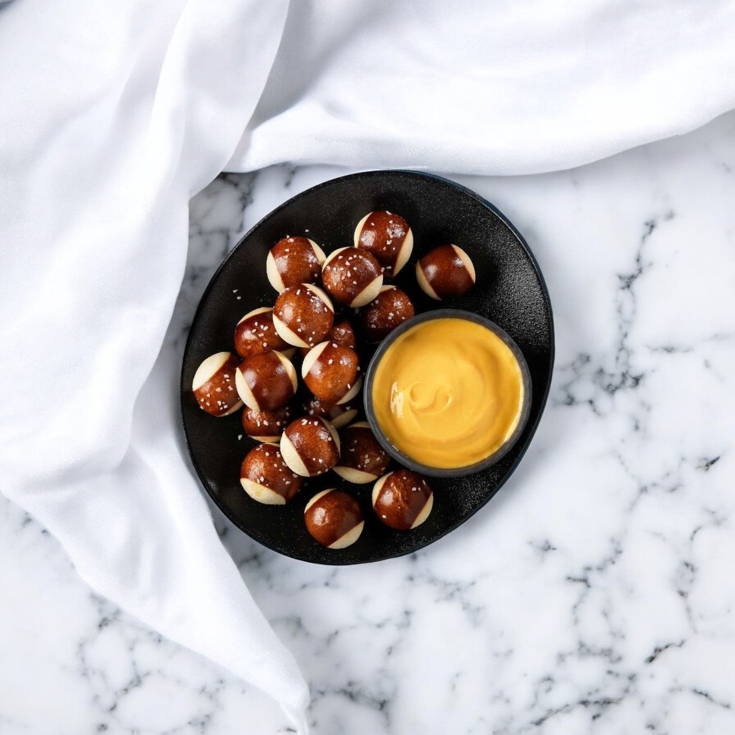 A black plate featuring Pretzilla Pretzel Bites, 36.5 oz. - 1 Box, is accompanied by a bowl of mustard sauce on a white marble surface, making it perfect for satisfying snack cravings. A white cloth is draped beside it, adding an elegant touch to this delicious setup.