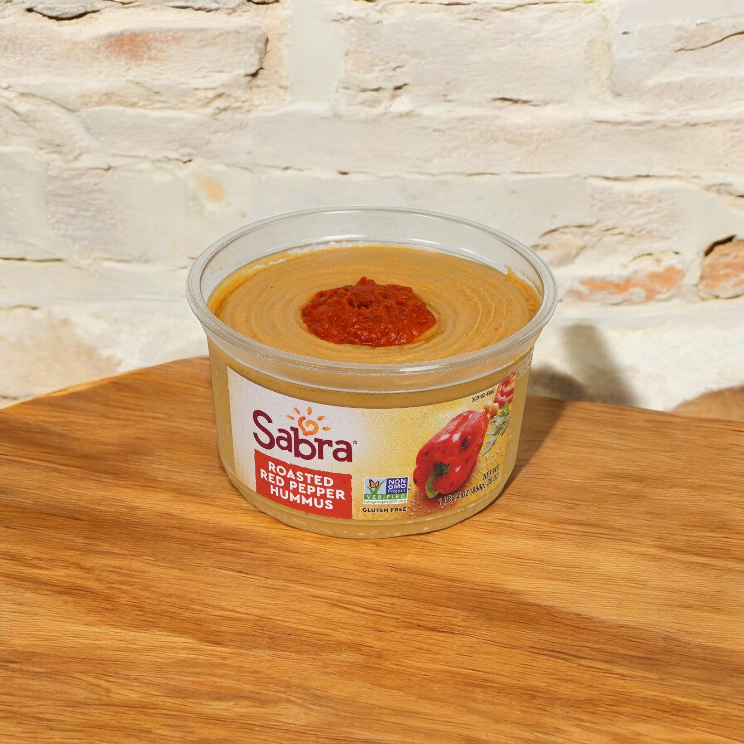 A container of Sabra Roasted Red Peppers Hummus Spread, 30 oz. - 1 Count, known for its creamy texture and natural ingredients, rests on a wooden surface against a light-colored brick backdrop.