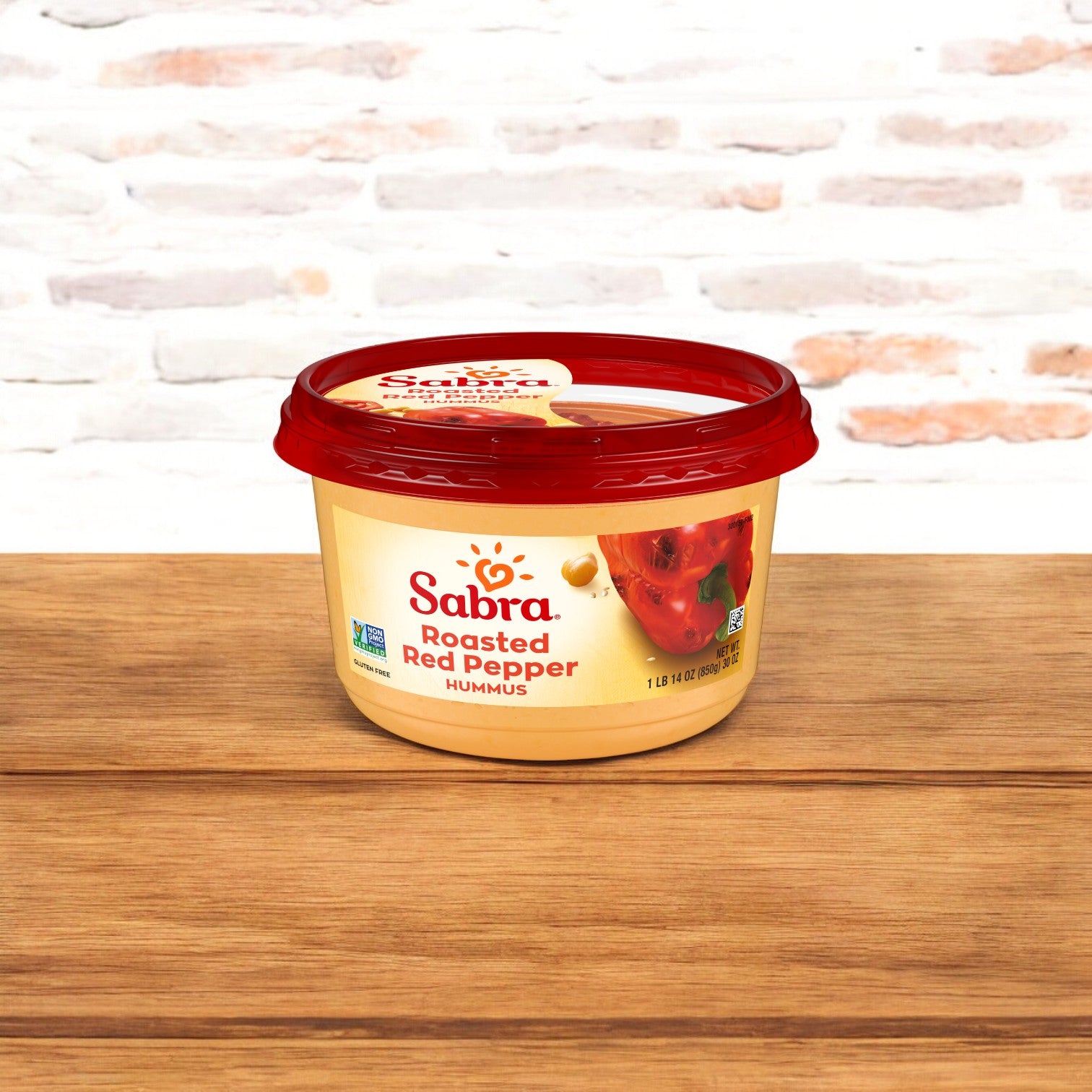 A container of Sabra Roasted Red Peppers Hummus Spread, 30 oz. - 1 Count, with a red lid and showcasing its creamy texture, sits on a wooden surface against a white brick background.