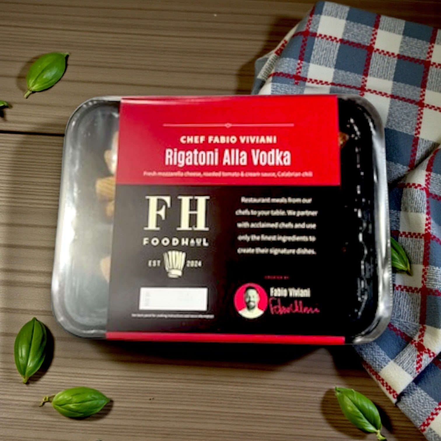FoodHaul Frozen Meal – FoodHaul Rigatoni Alla Vodka by Chef Fabio Viviani sits on a wooden surface, accompanied by scattered basil leaves and a checkered cloth.