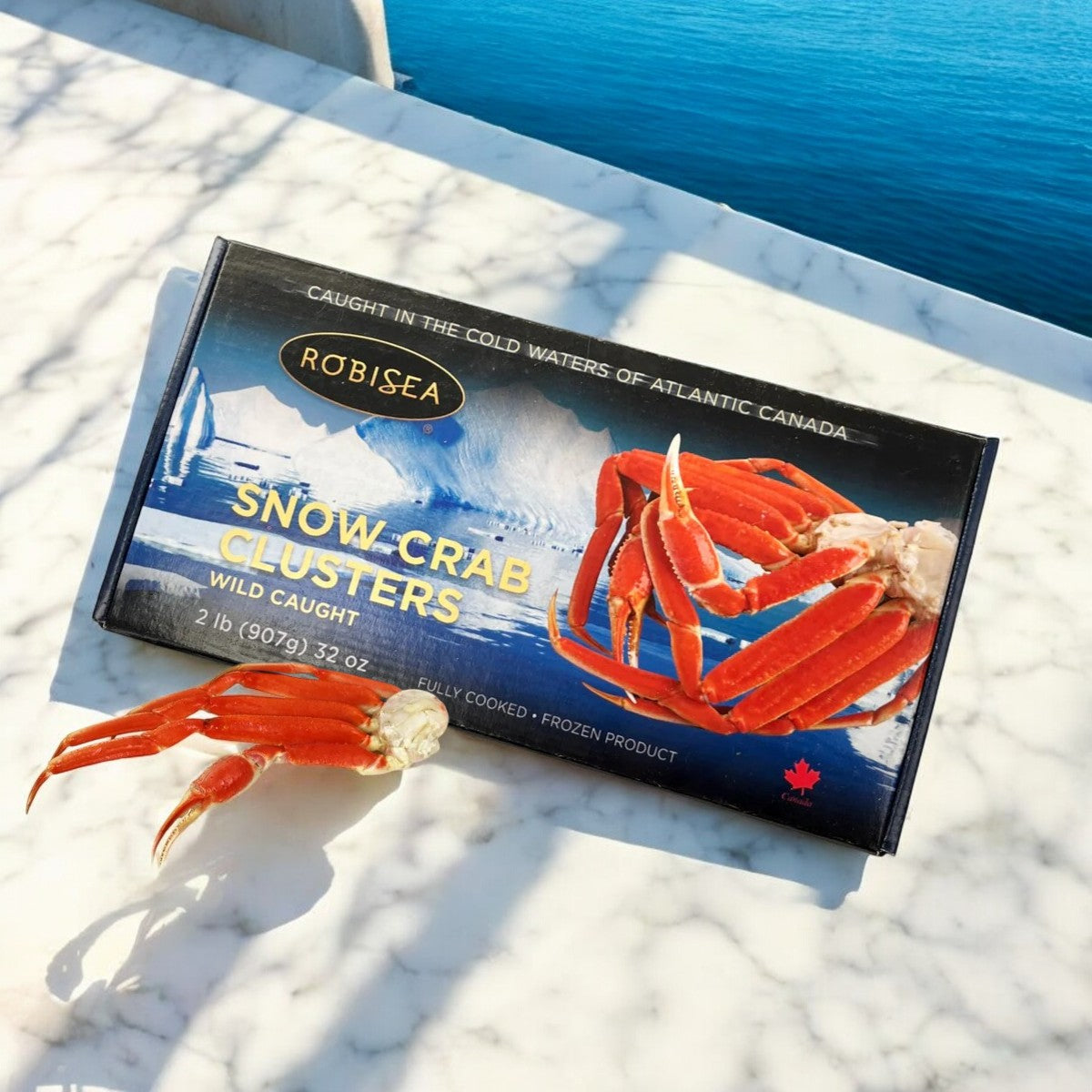 On a marble surface lies a box of Easy Lunches' Robisea Wild Caught Snow Crab Clusters, 2 lb., complemented by a lobster tail artfully placed next to a crab claw. In the background, blue water and a railing create an ideal ambiance for an exquisite surf & turf dining experience.