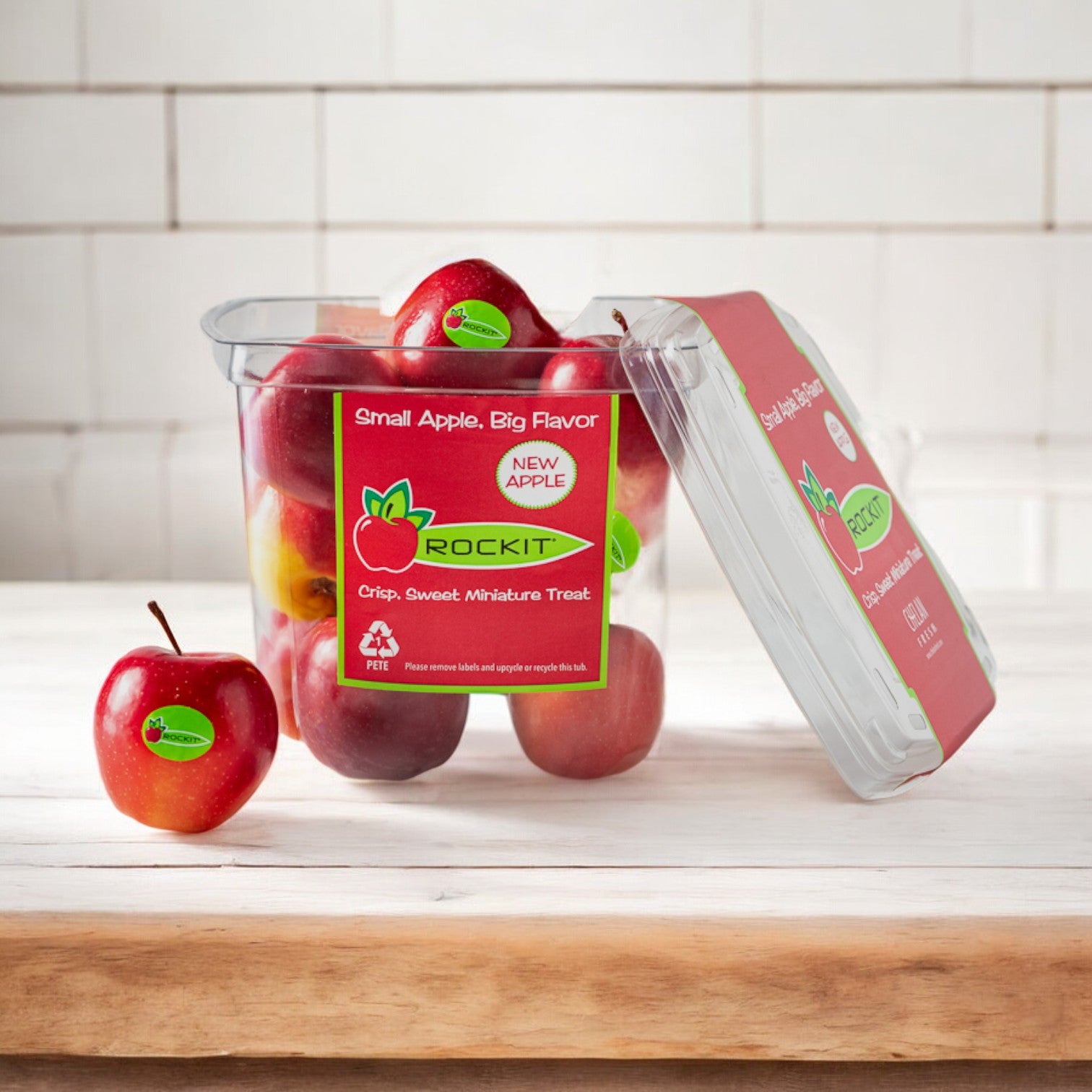 A clear container of Rockit Crisp Sweet Miniature Apples by Easy Lunches, with one small red apple outside the container, sits on a wooden counter in a kitchen with white tiled walls. The label reads "Small Apple, Big Flavor.