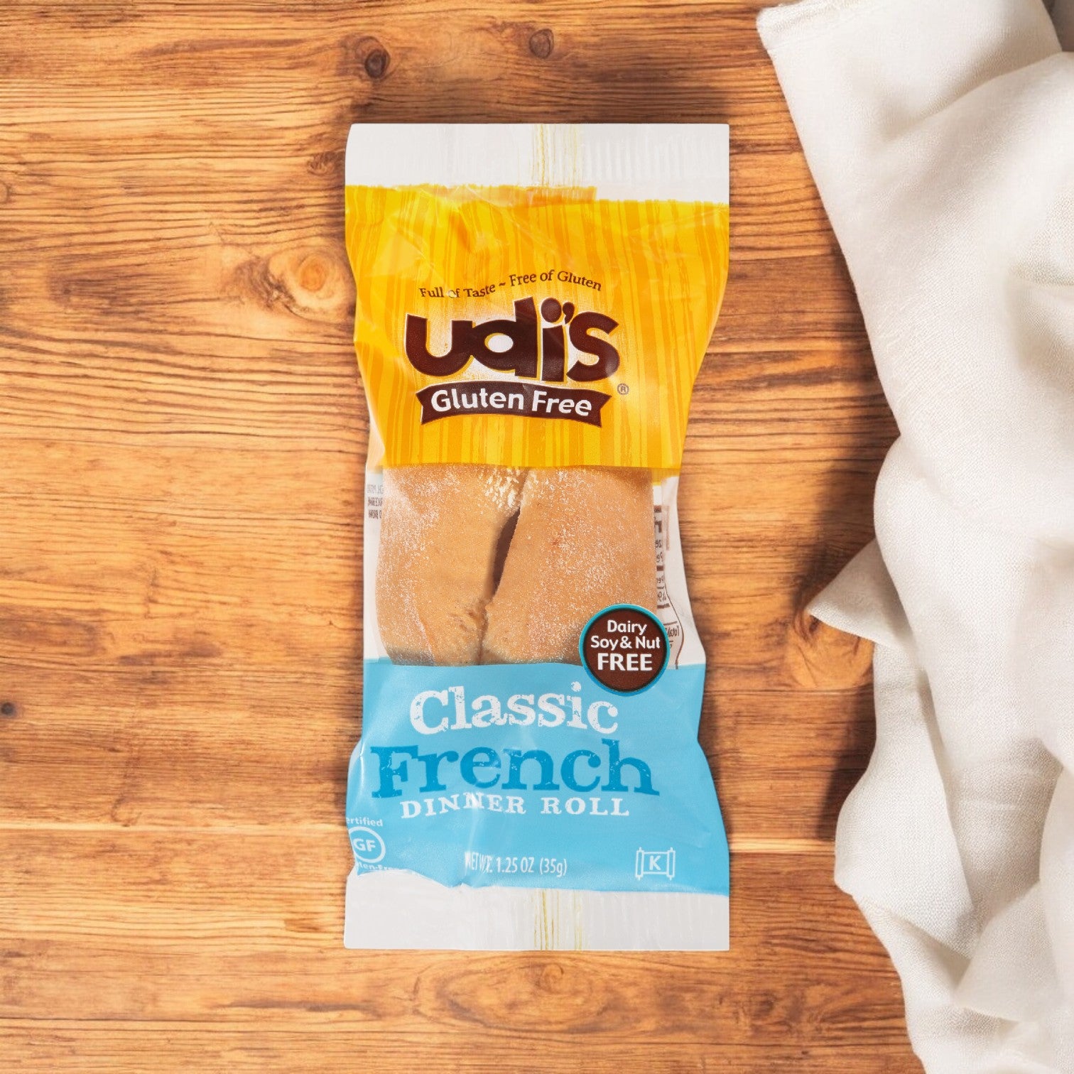 A package of Udi's Roll Dinner French Gluten Free sits on a wooden surface with its soft and fluffy texture, accompanied by a nearby white cloth.