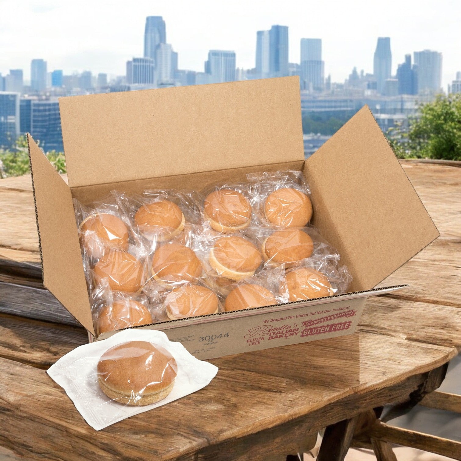 Open a Rotella Bun Hamburger Vegan Gluten Free-24 Count box on a wooden table with individually wrapped buns, city skyline in the background.