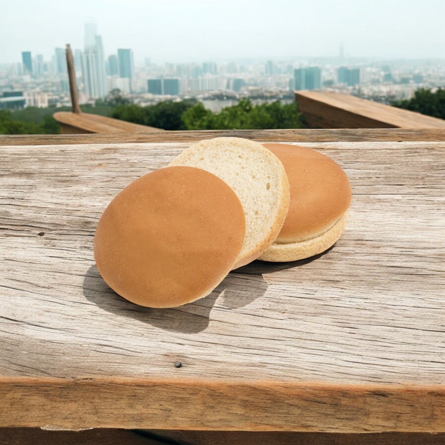 Two Rotella Bun Hamburger Vegan Gluten Free buns sit on a wooden surface, framed by a cityscape in the background.