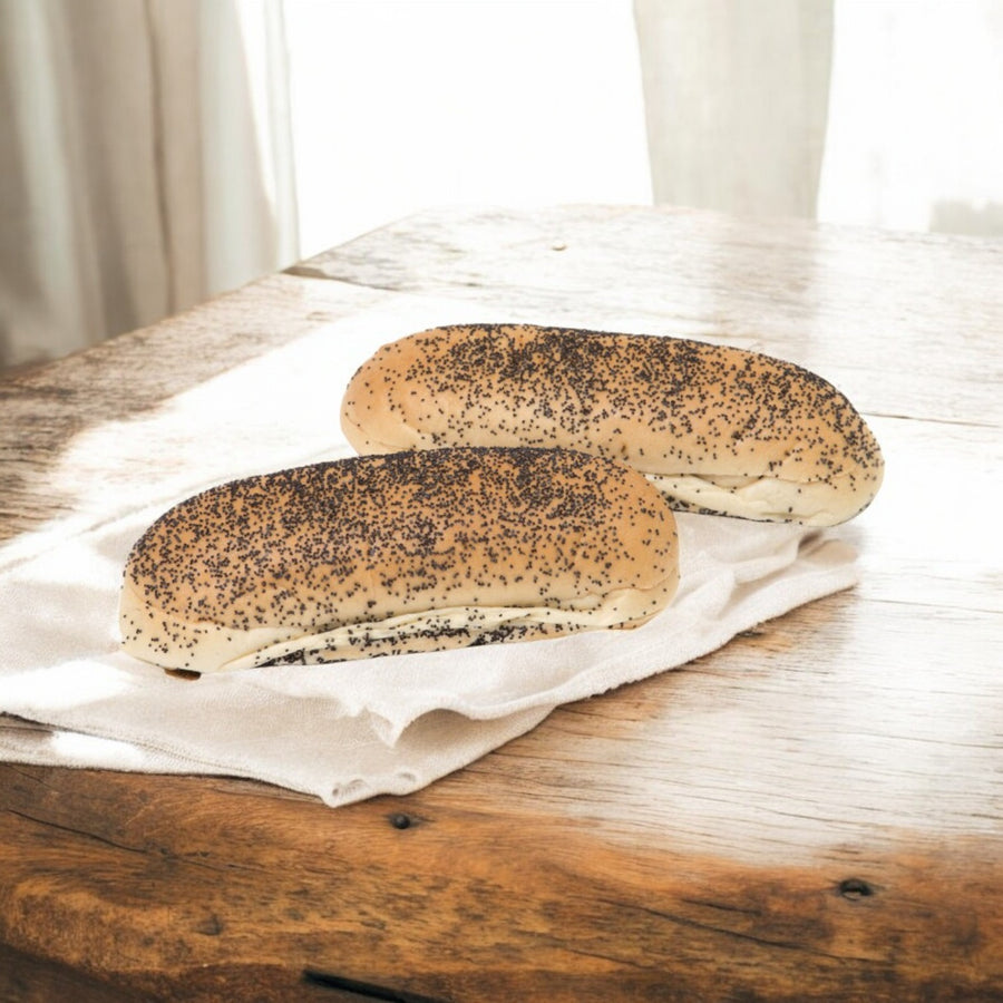 Two Rotellas Poppyseed Sliced Hot Dog Buns from Easy Lunches rest on a white cloth on a sunlit wooden table. These 5.5" artisanal delights, reminiscent of soft deli buns, are ready to elevate any meal.