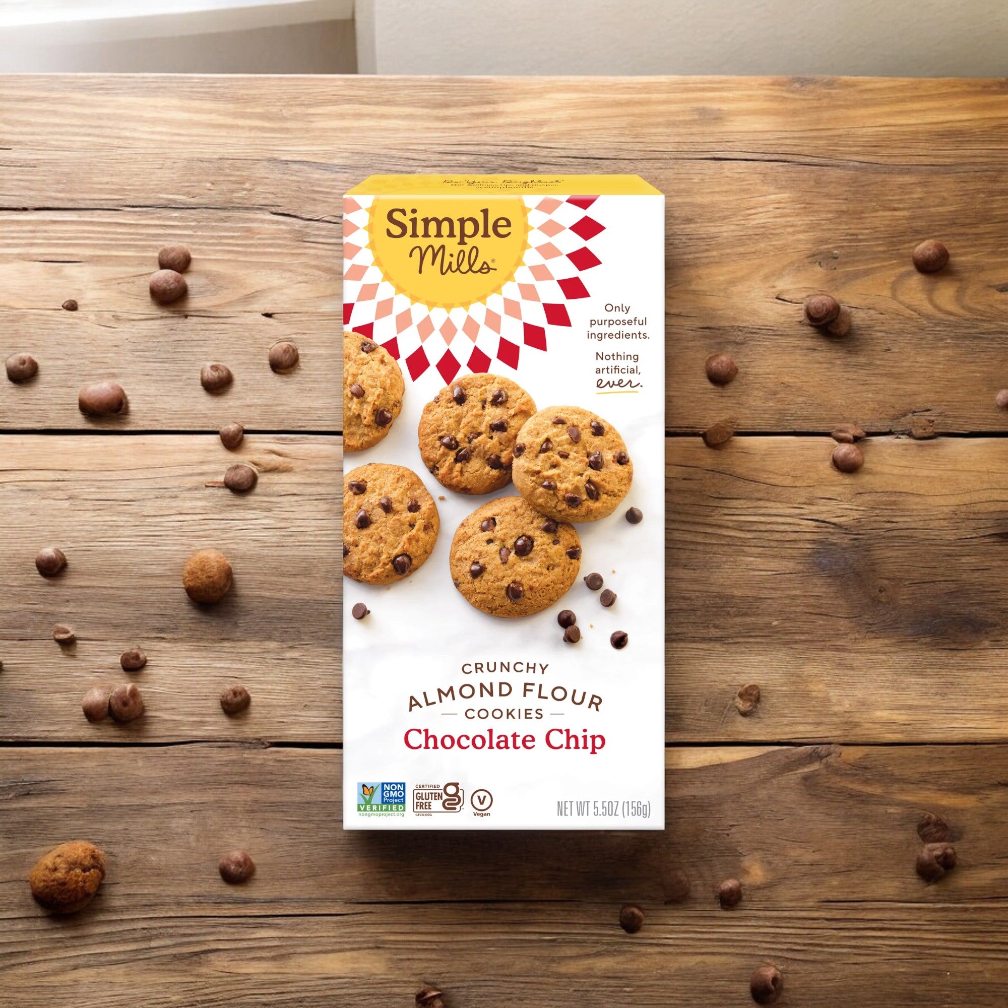 A box of Simple Mills Chocolate Chip Cookies is placed on a wooden surface, highlighting their all-natural ingredients. Several gluten-free cookies and chocolate chips are scattered around the 5.5 oz Simple Mills box, tempting you to indulge.