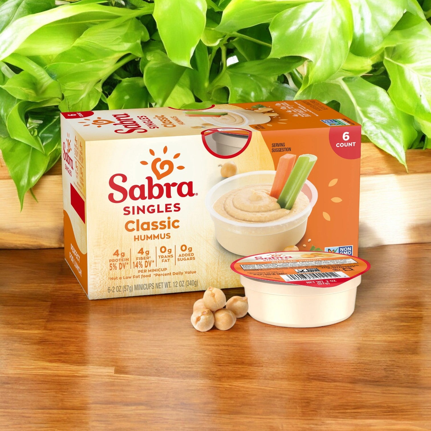 A box of Sabra Classic Hummus Singles with one individual container in front, set against a leafy background. This plant-based snack from Sabra contains six 2 oz servings and is labeled gluten-free, kosher, and non-GMO. Enjoy convenient single-serve cups perfect for on-the-go snacking.