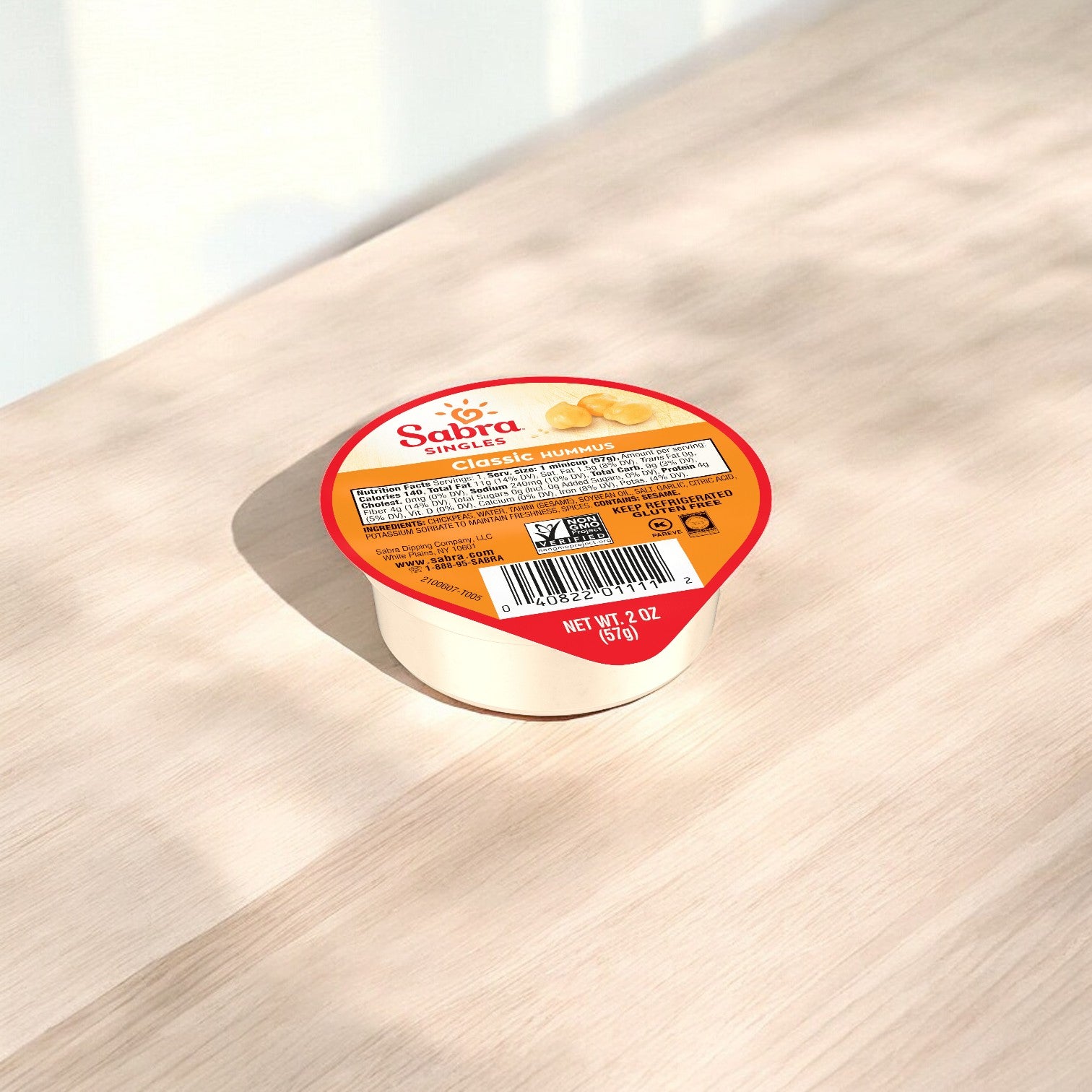 A single-serve container of Sabra Classic Hummus Singles, 2 oz Plastic Cups - 1 Count, ideal for a portable snack, rests on a light wooden surface. The packaging showcases nutritional information along with the Non-GMO Project Verified label.