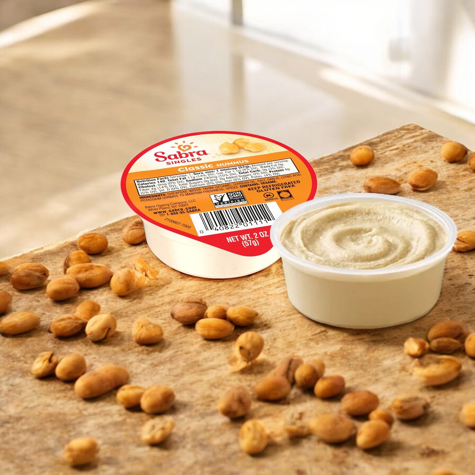 A container of Sabra Classic Hummus Singles, 2 oz Plastic Cups - 1 Count, is shown open on a wooden countertop, surrounded by scattered peanuts. Perfect as a single-serving hummus, it's an ideal portable snack for any time of the day.