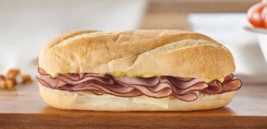 An Easy Lunches sub sandwich from the Sandwich Variety Pack, featuring layers of sliced smoked ham, Swiss cheese, and mustard on a baguette-style roll, placed on a wooden surface.