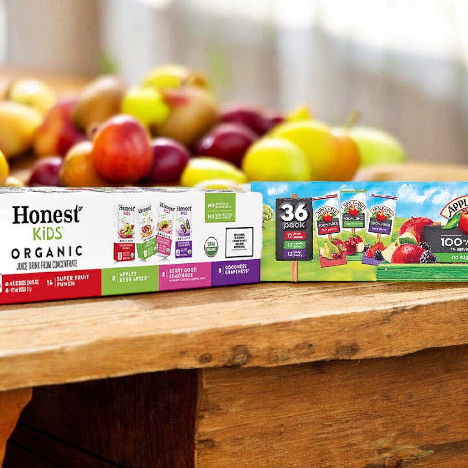 A carton of Easy Lunches Kid's Juice Pack, containing 40 assorted Honest Kids juice boxes, along with two packs of 36 assorted kids' juice boxes each, are placed on a wooden surface with a variety of fruits in the background. These beverages boast reduced sugar and organic ingredients for a healthier choice.