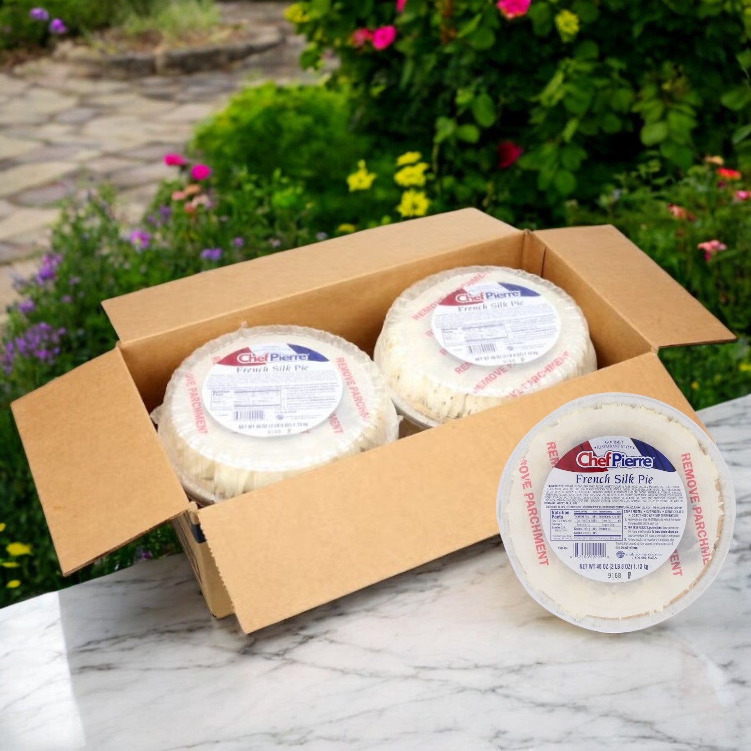 An open cardboard box containing four Chef Pierre 10-inch Chocolate French Silk Pies, each with a rich chocolate filling, is set on a marble surface against a lush garden backdrop.