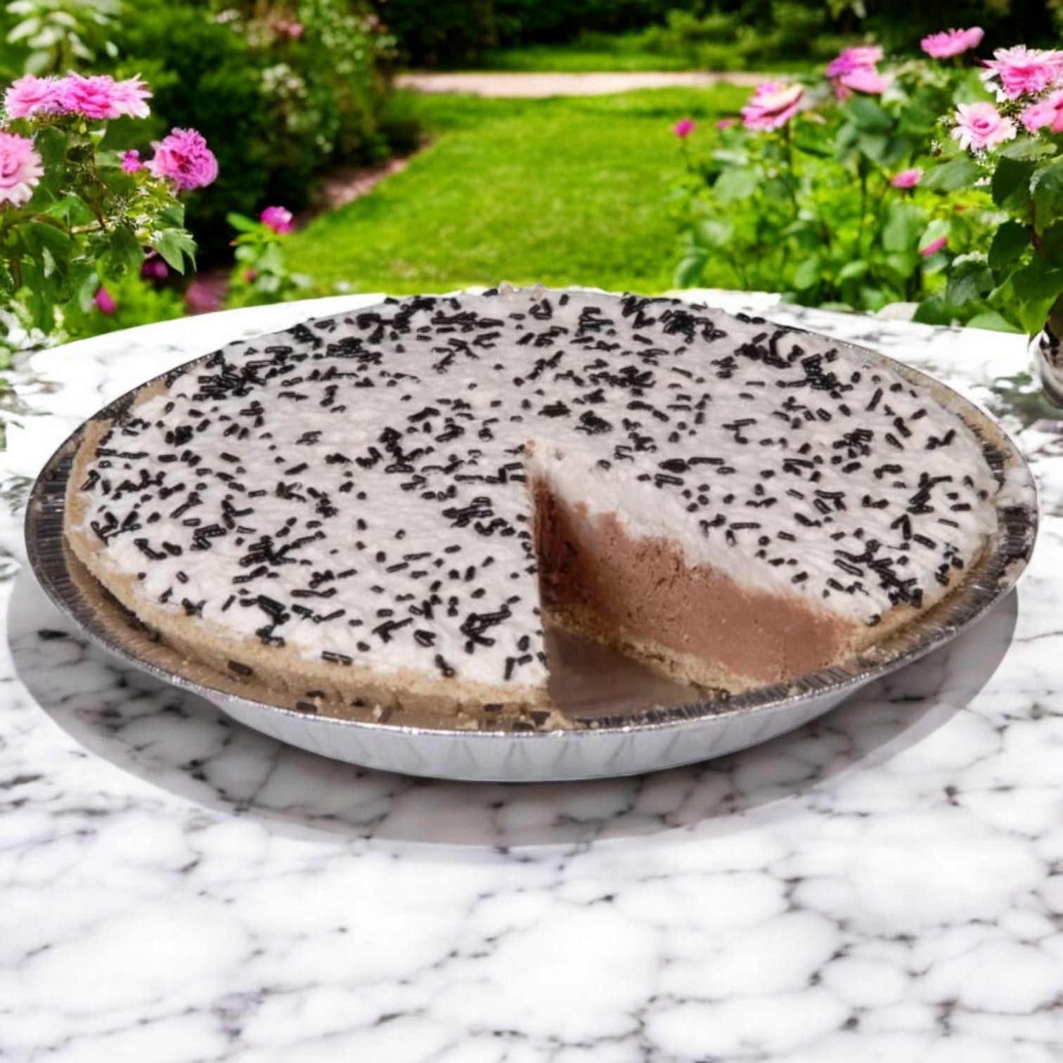 A Chef Pierre Chocolate Cream Pie with a slice removed rests as a stunning 10-inch frozen dessert on a marble table, surrounded by pink flowers and lush greenery outdoors, topped with sprinkles.