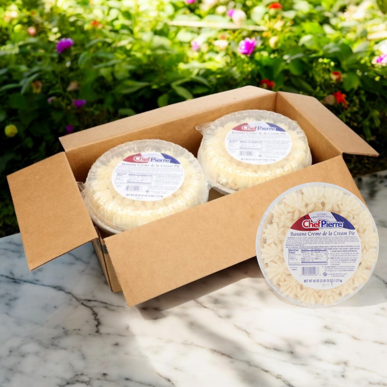 A cardboard box holding four Chef Pierre Banana Crème de la Cream 10-Inch Frozen Pies rests on a marble surface amid lush greenery and flowers. The creamy banana filling tantalizingly peeks out, promising indulgence in every bite.