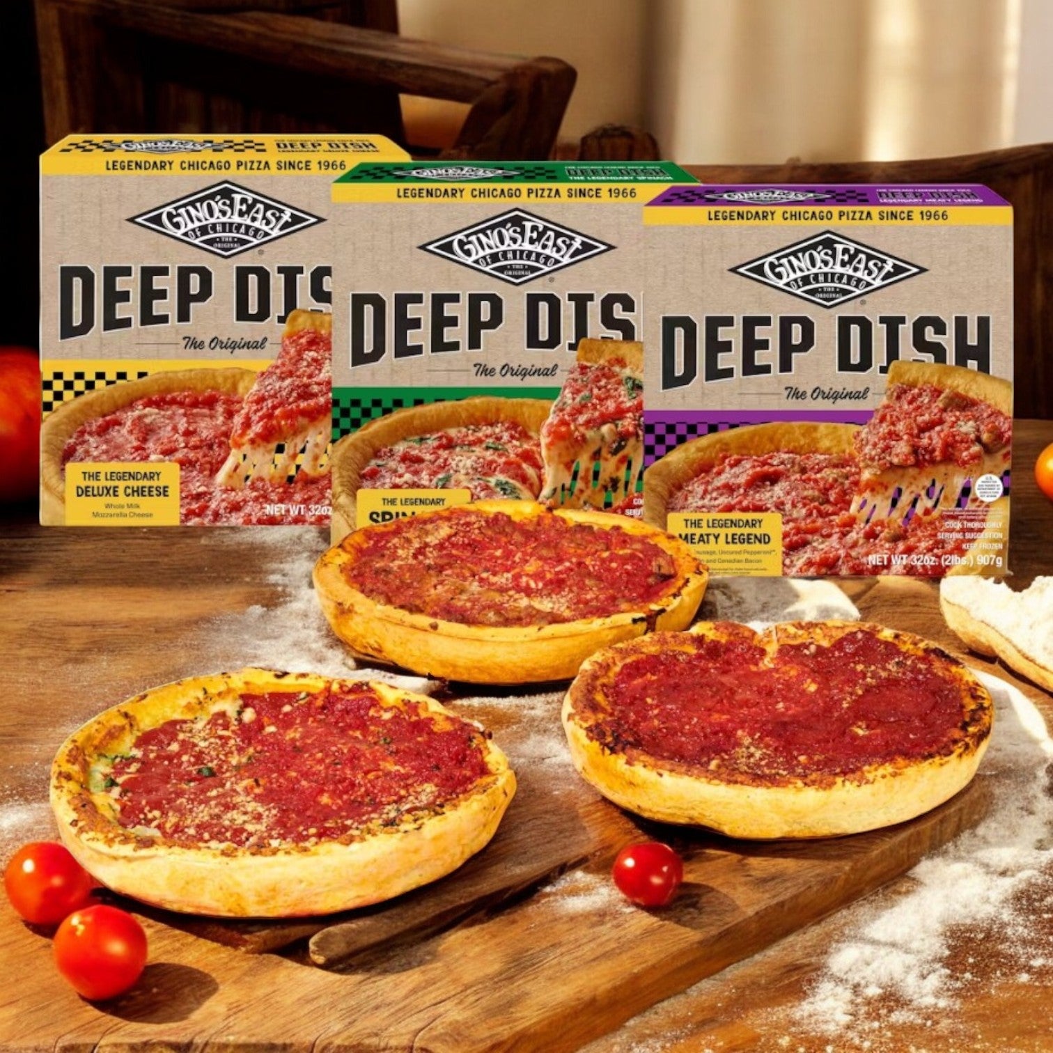 Three deep-dish pizzas—Spinach Deep Dish, The Legendary Meaty Legend, and The Legendary Deluxe Cheese—are displayed in front of Gino's East Pizza boxes on a wooden table, surrounded by ripe tomatoes.