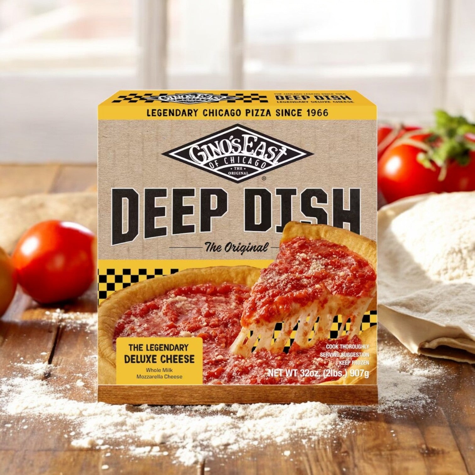 A Gino's East Pizza box from their 3-Pack Combo (Spinach Deep Dish, Meaty Legend, Deluxe Cheese) sits on a wooden table surrounded by tomatoes, dough, and flour. The tantalizing image on the box features their iconic cheesy deep-dish slice.