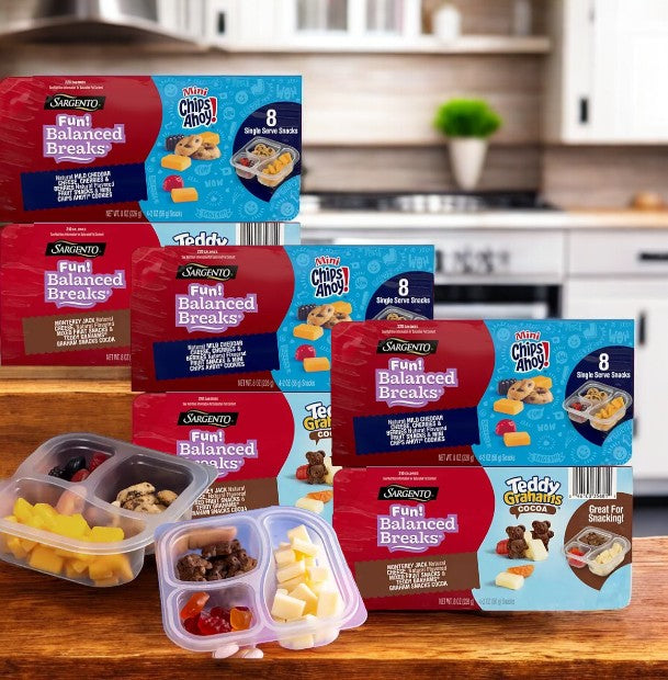 Several boxes of snacks, featuring Sargento Fun Balanced Breaks Variety Pack with 24 packs, Chips Ahoy, Teddy Grahams, and other treats are displayed on a kitchen counter alongside fruit and cheese in containers.