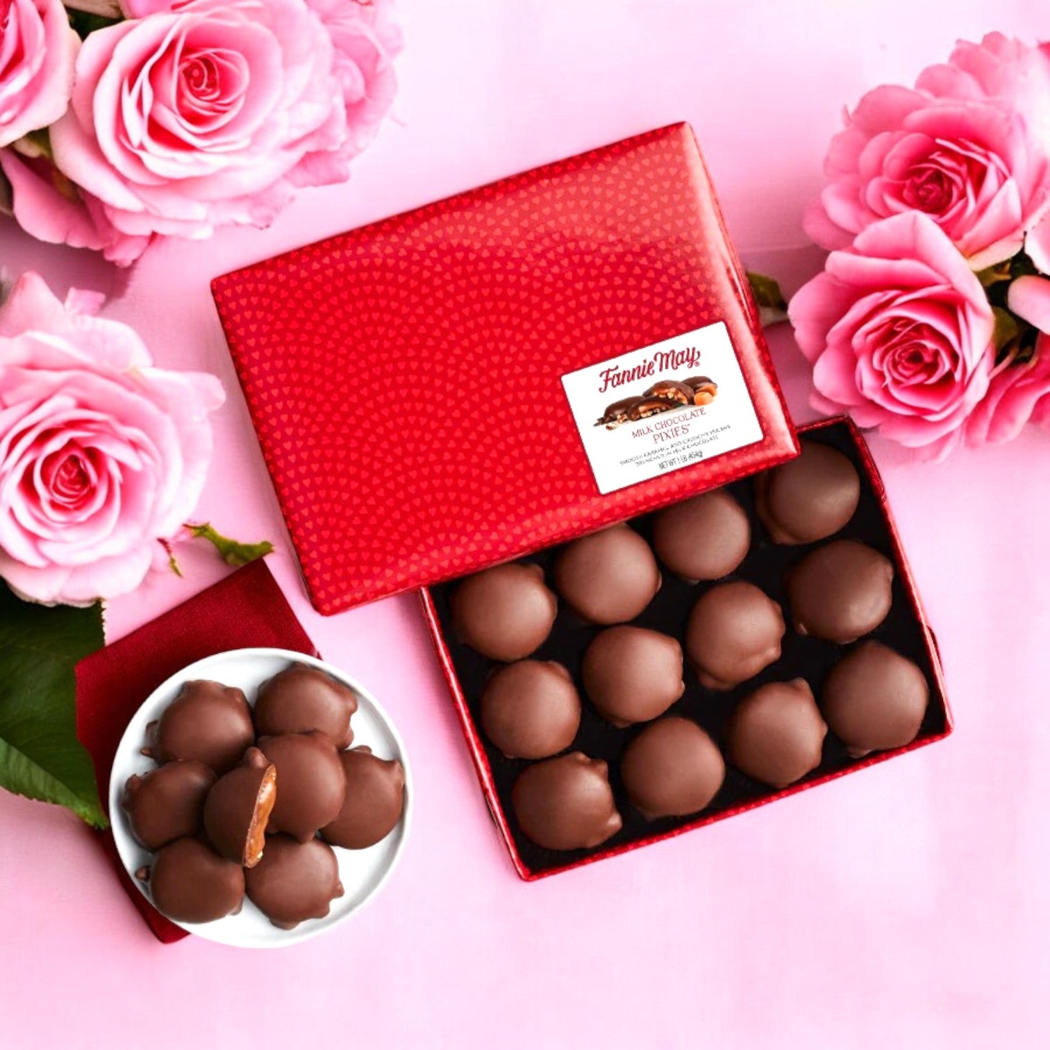 A red box of The Garrett Mix and Fannie May Pixel Combo by Easy Lunches sits open beside a bowl with three more chocolates. Pink roses are artfully scattered across a light pink surface, crafting an enchanting scene reminiscent of the perfect sweet and savory treat.