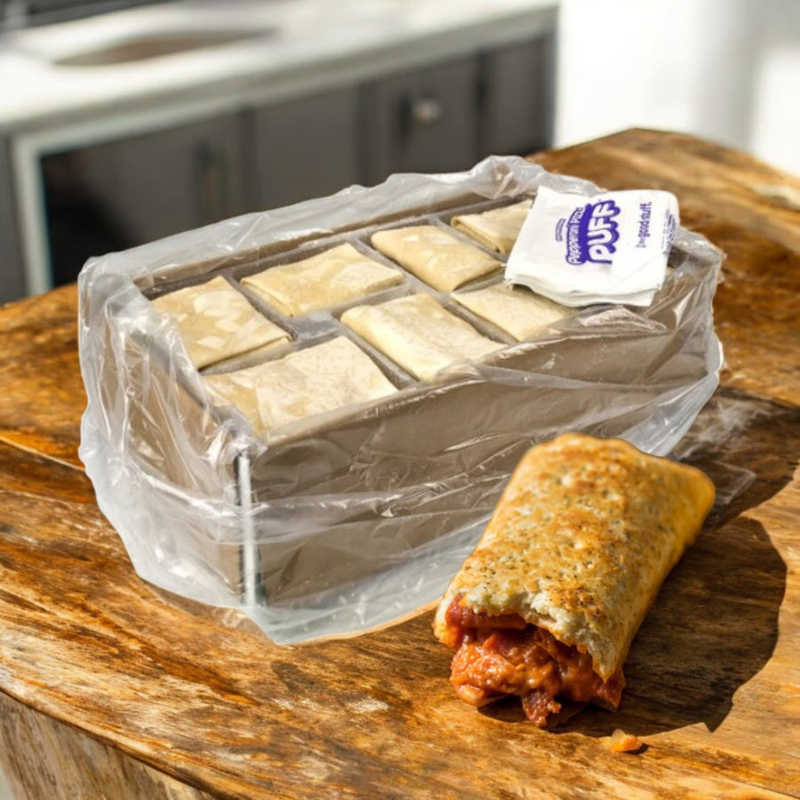 A case of Iltaco Foods Italco Puff Pepperoni Pizza (48 count, 6 oz each) in plastic packaging is displayed on a wooden surface, with one cooked pastry bitten open to show its red pepperoni filling.