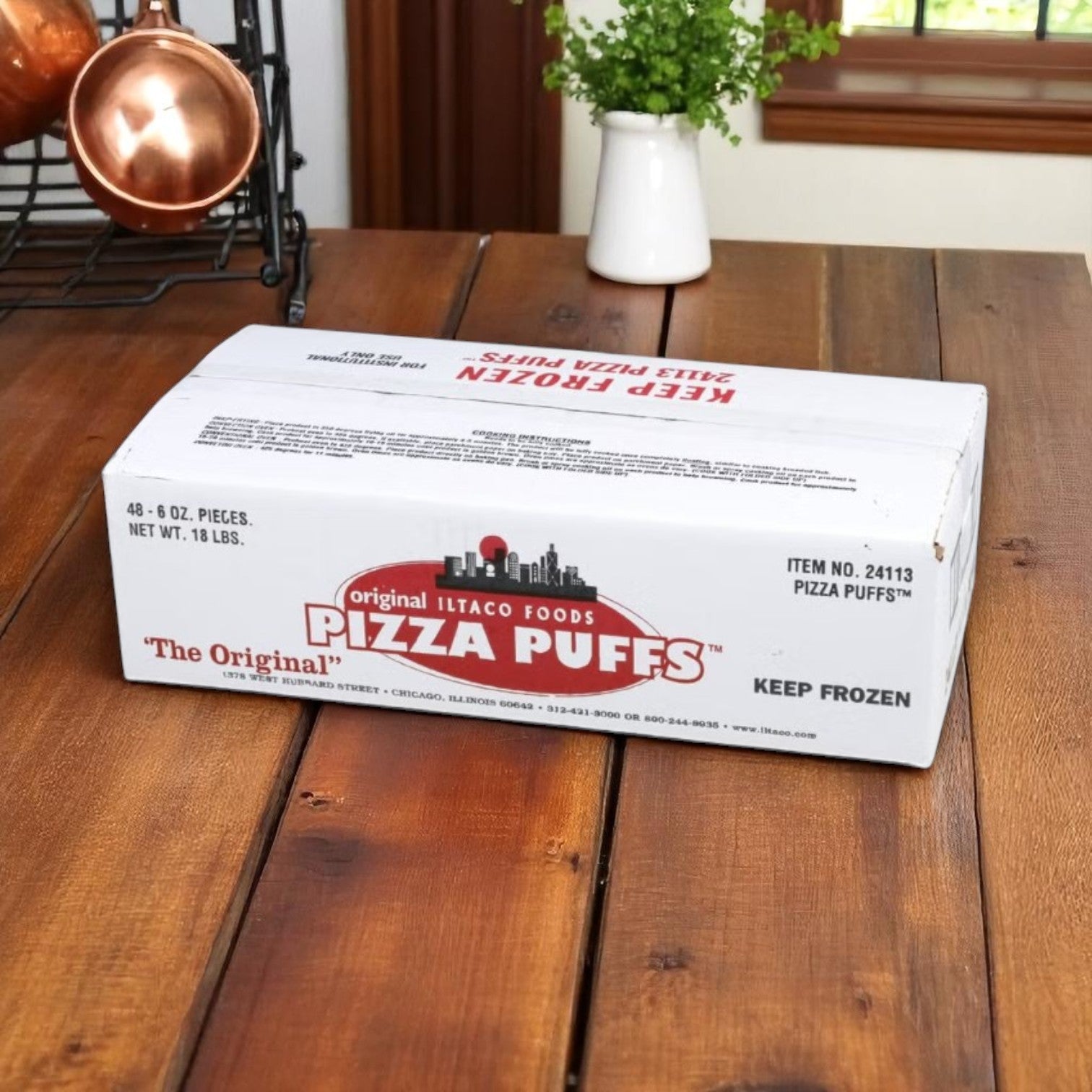 A white box labeled "The Original Pizza Puff" sits on a wooden kitchen table, featuring Iltaco Sausage & Cheese Pizza Puff – 6 oz (48 per case), near copper pans and a small plant by the window.