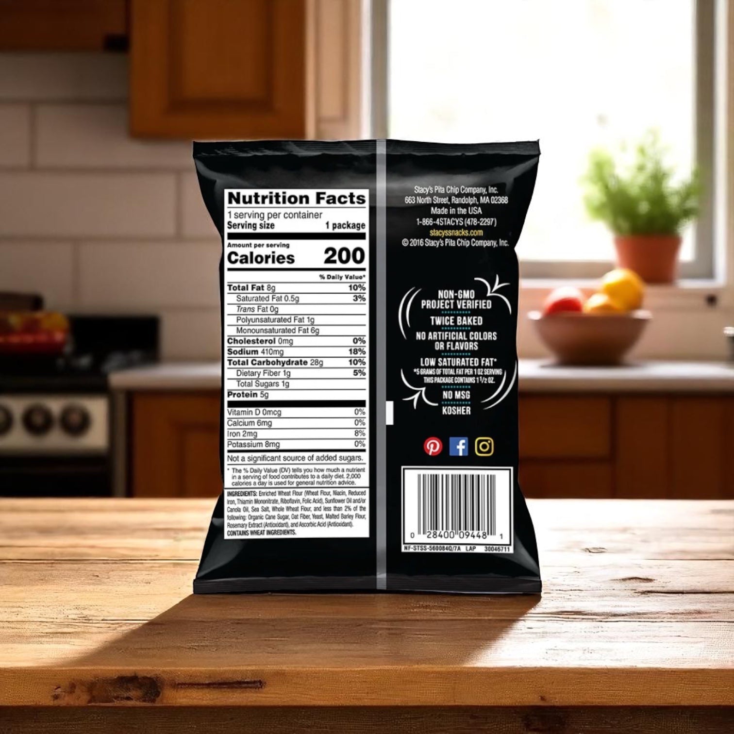 A bag of Easy Lunches Stacys Pita Chips Large Single Serving Simply Naked (24 case) stands upright on a kitchen counter, displaying its nutrition label and barcode, with a blurred kitchen and fruit bowl in the background—ready to enjoy.