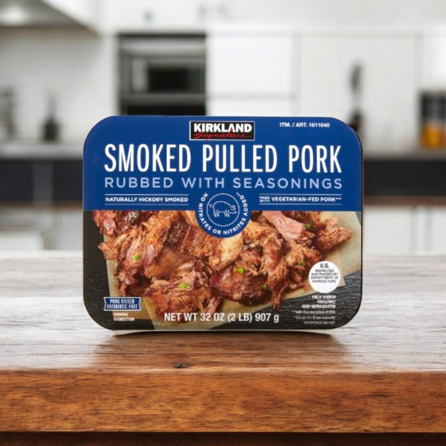 A 32 oz container of Kirkland Signature Smoked Pulled Pork, rubbed with seasonings, sits on a wooden kitchen counter.